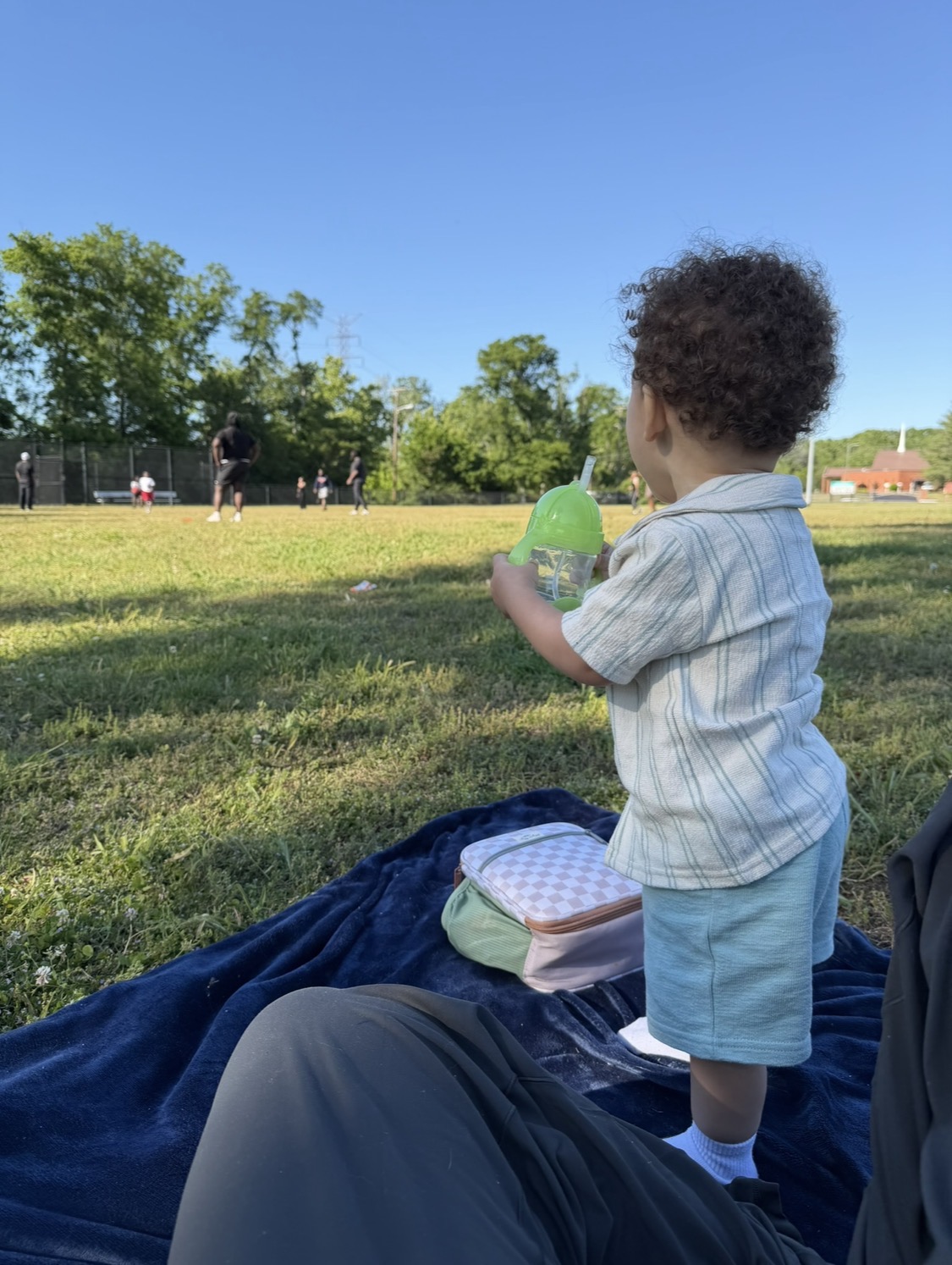Child standing on a blanket in a park holding a green bottle, with trees and people in the background.