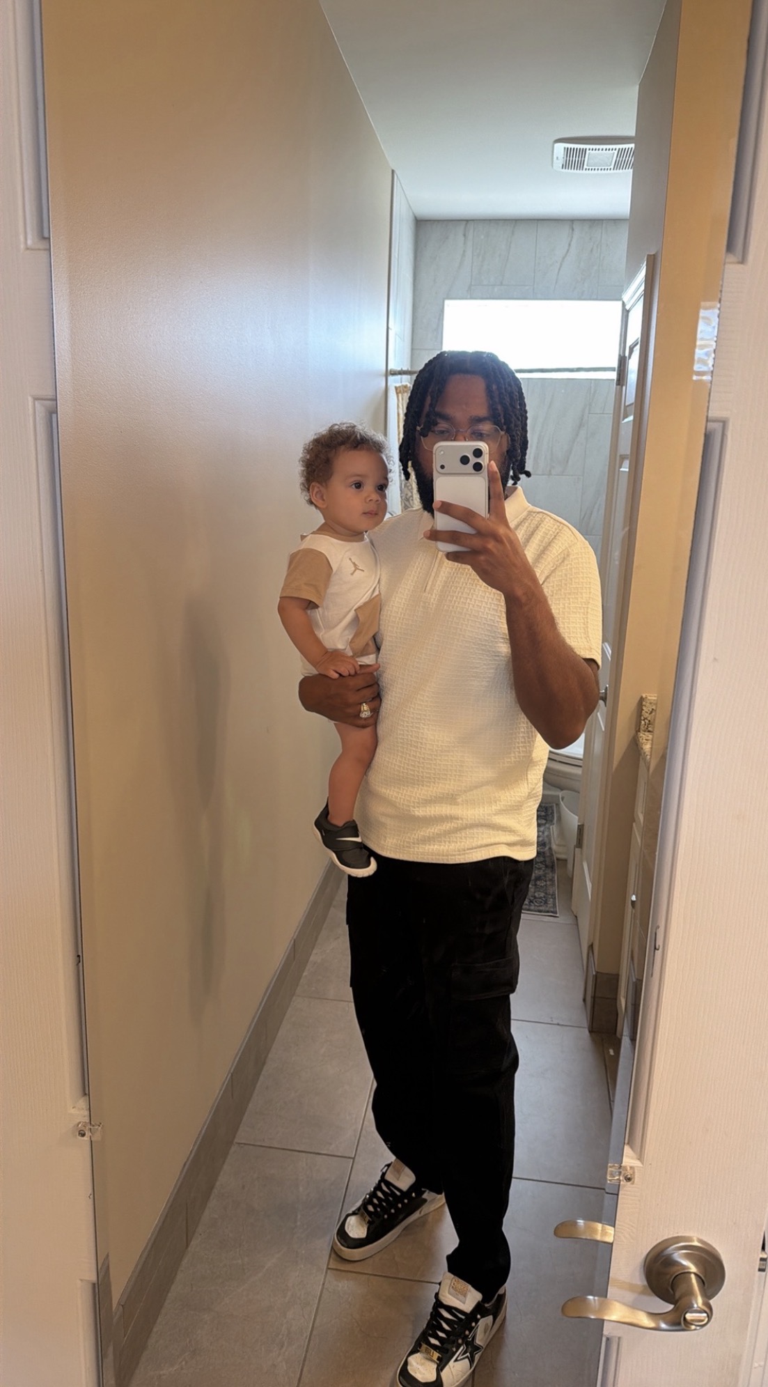 Person holding a child in a hallway, taking a mirror selfie, with a door and window in the background.