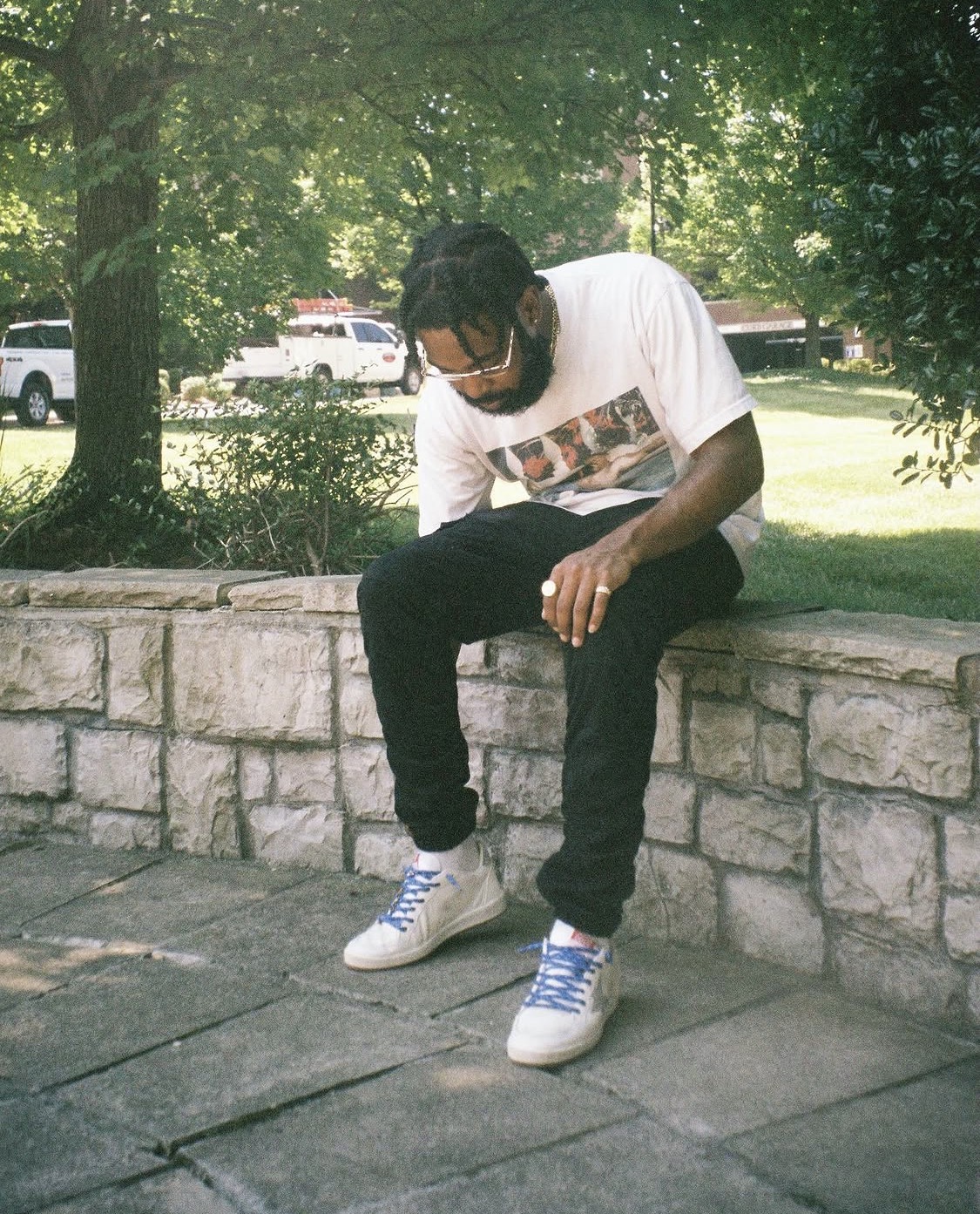 Person sitting on a stone ledge outdoors, wearing a white t-shirt, black pants, and white sneakers, with trees and parked cars in background.