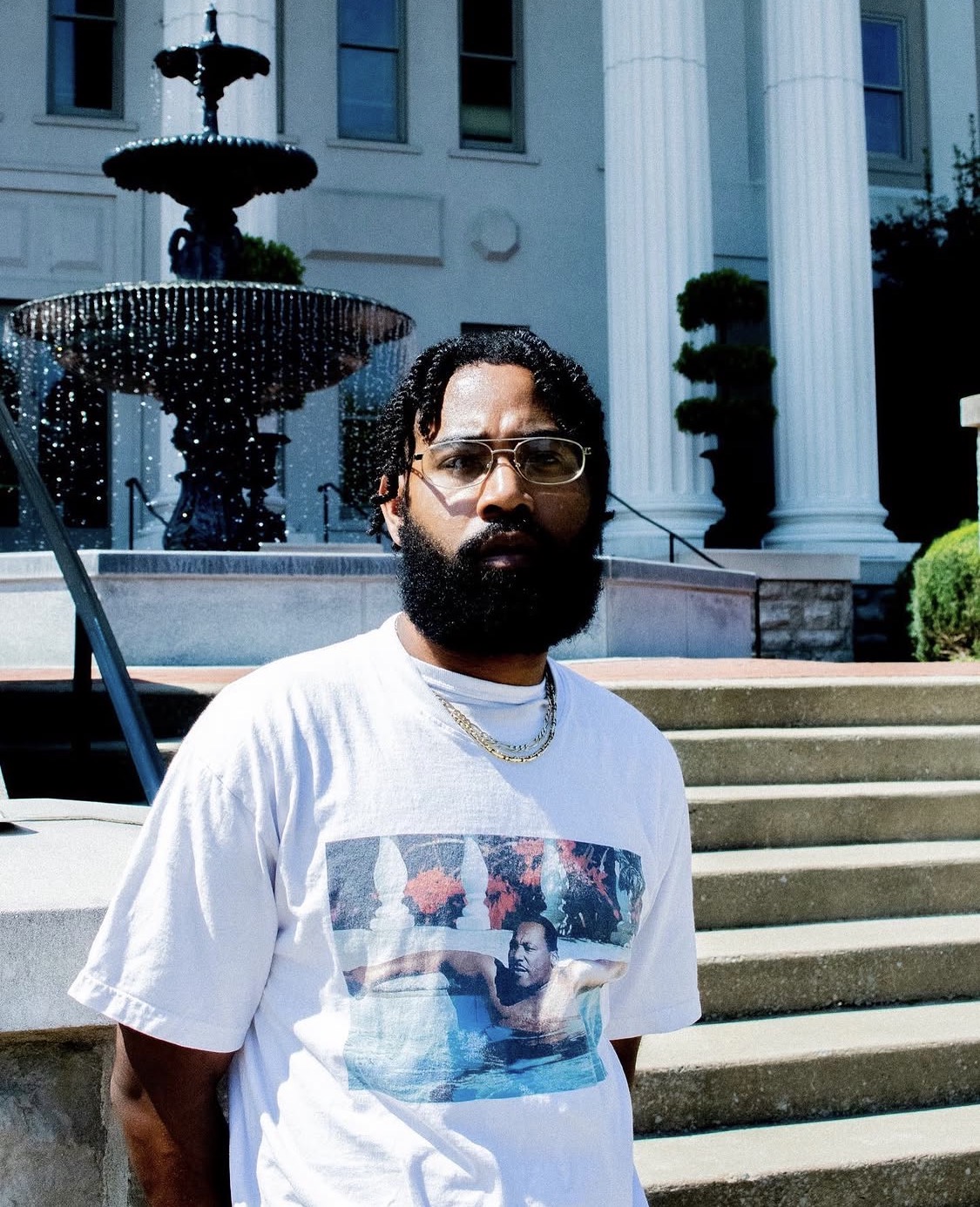 Man with glasses and a beard stands outdoors in front of a fountain and building, wearing a white T-shirt with a graphic.