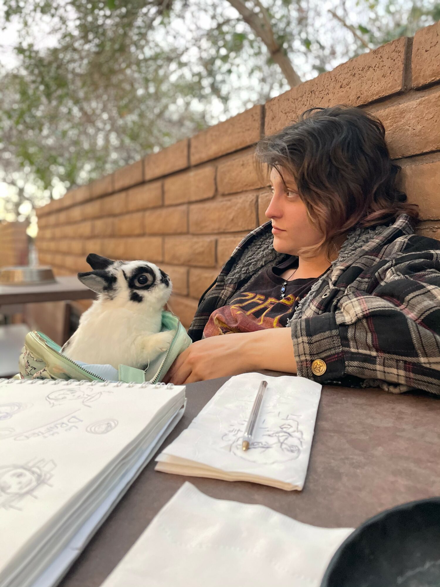 Person with short dark hair and plaid shirt sitting at table with a black and white cat, sketchbooks and pens present.