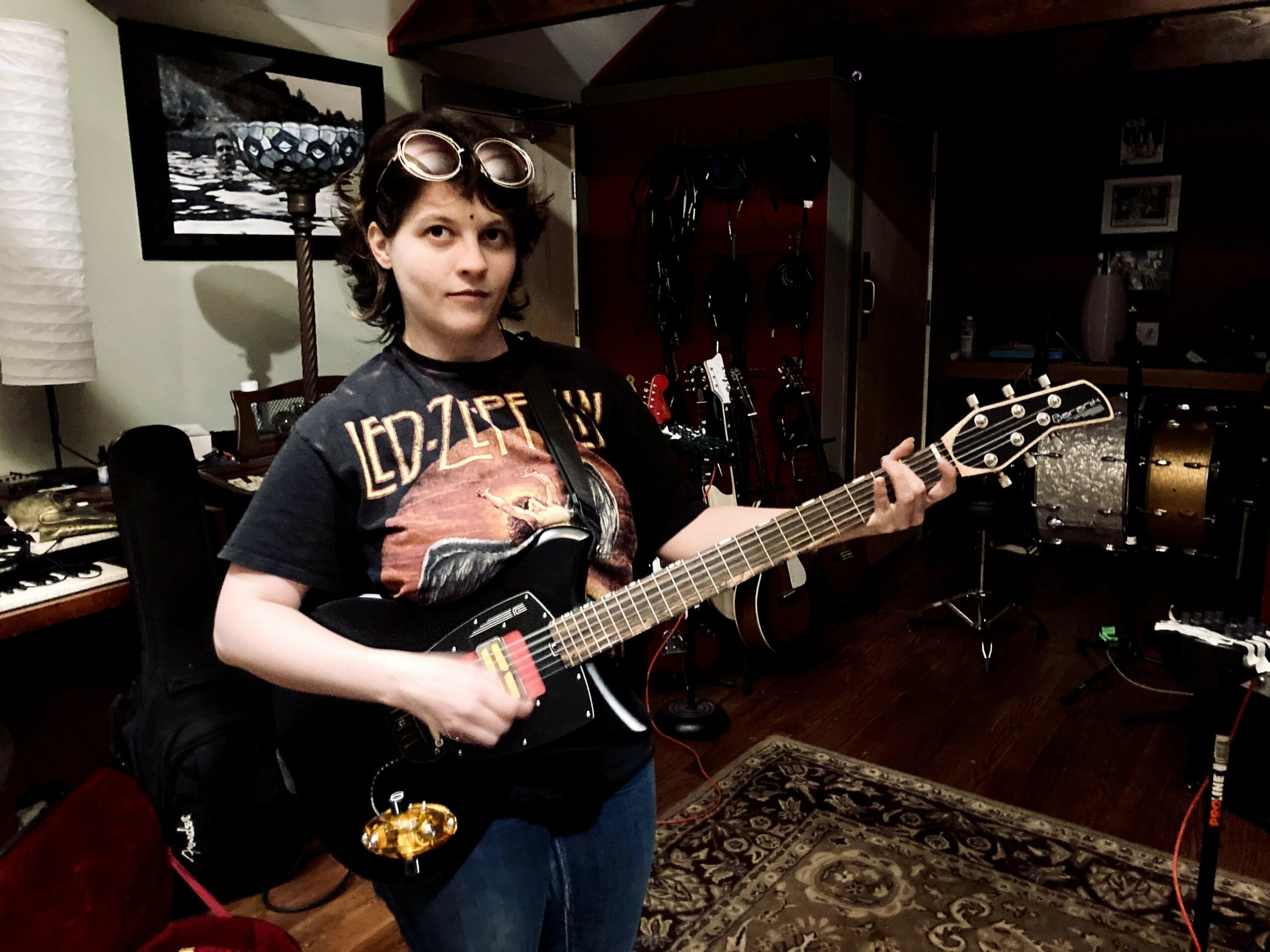 Person holding a guitar in a room with musical instruments and furniture, wearing a Led Zeppelin T-shirt and sunglasses on head.