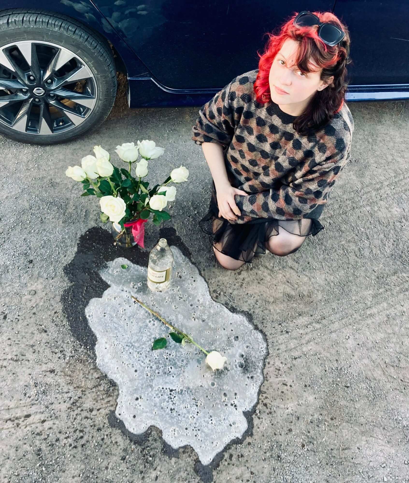 Woman with red hair and sunglasses kneels near a spilled liquid, white roses, and a bottle on the ground beside a car.