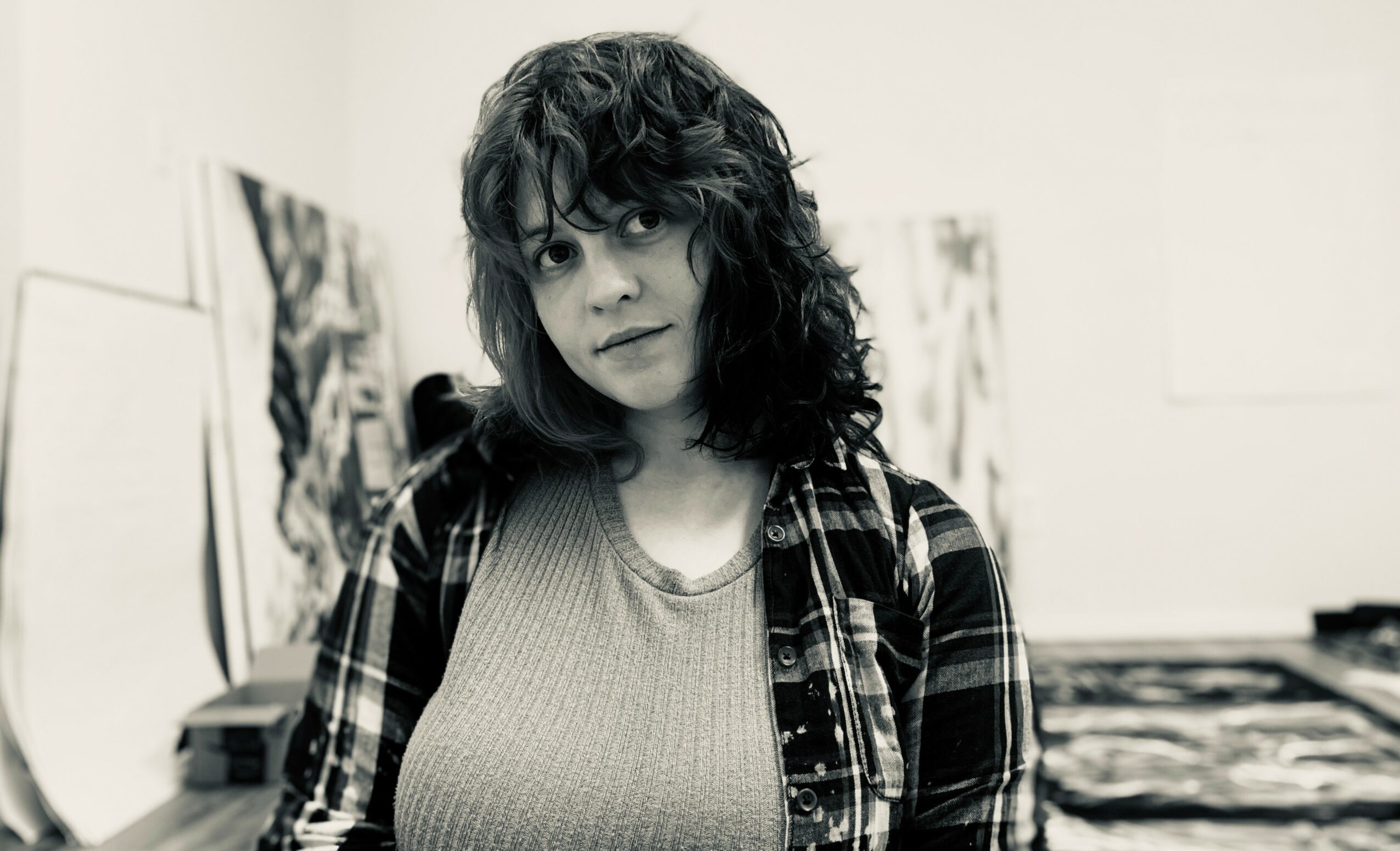 Young woman with curly hair wearing a plaid shirt over a top, standing in an art studio with paintings in the background.