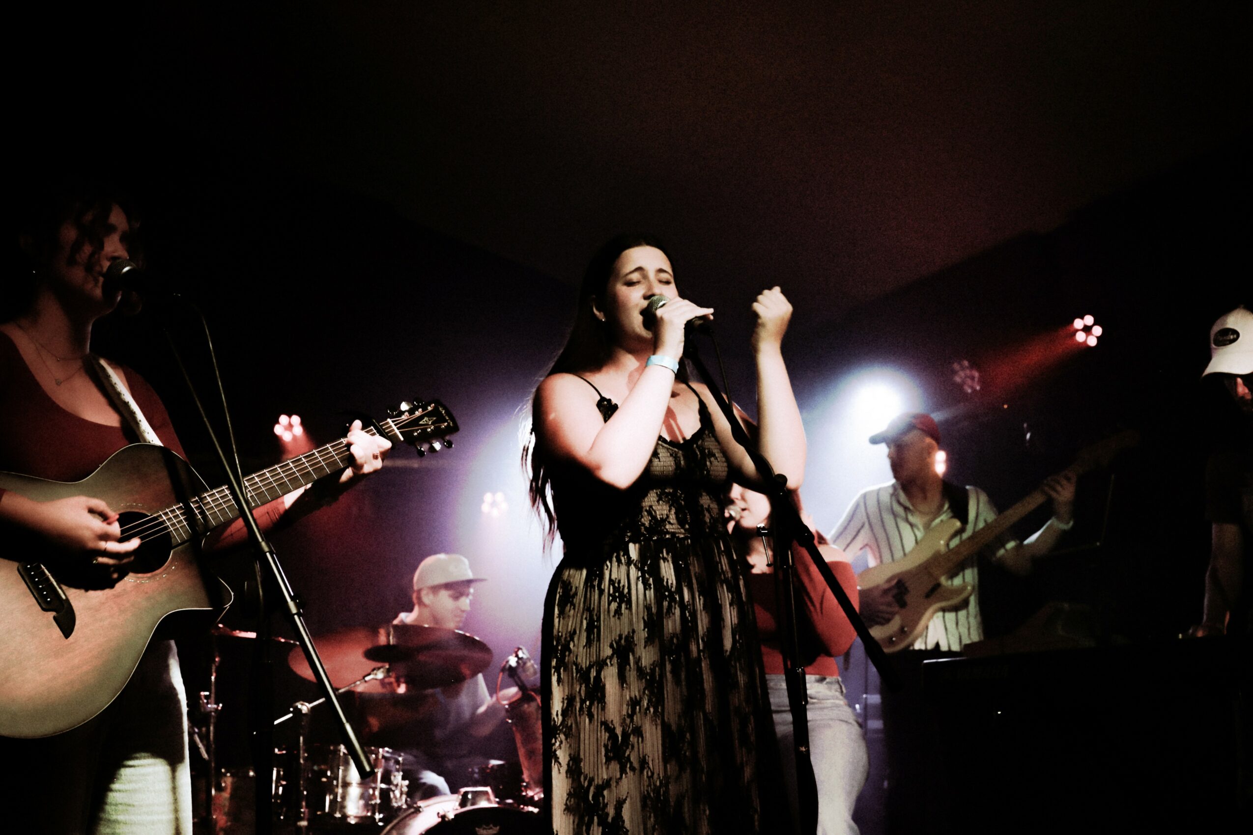 Group of musicians performing on stage, with a woman singing into a microphone, surrounded by guitarists and a drummer, dim lighting.