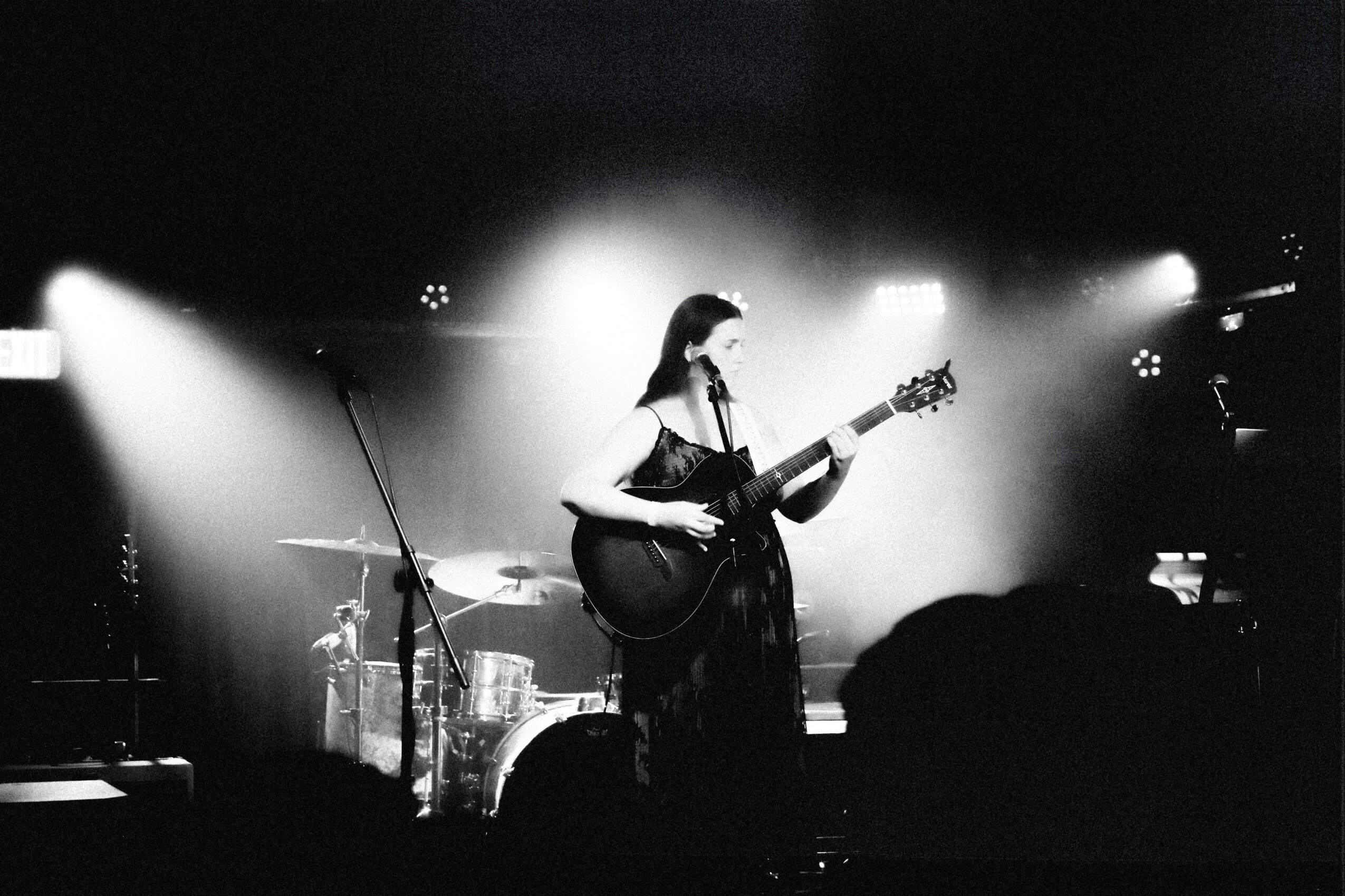 A woman plays guitar on stage with drums and bright lights behind her, in black and white.