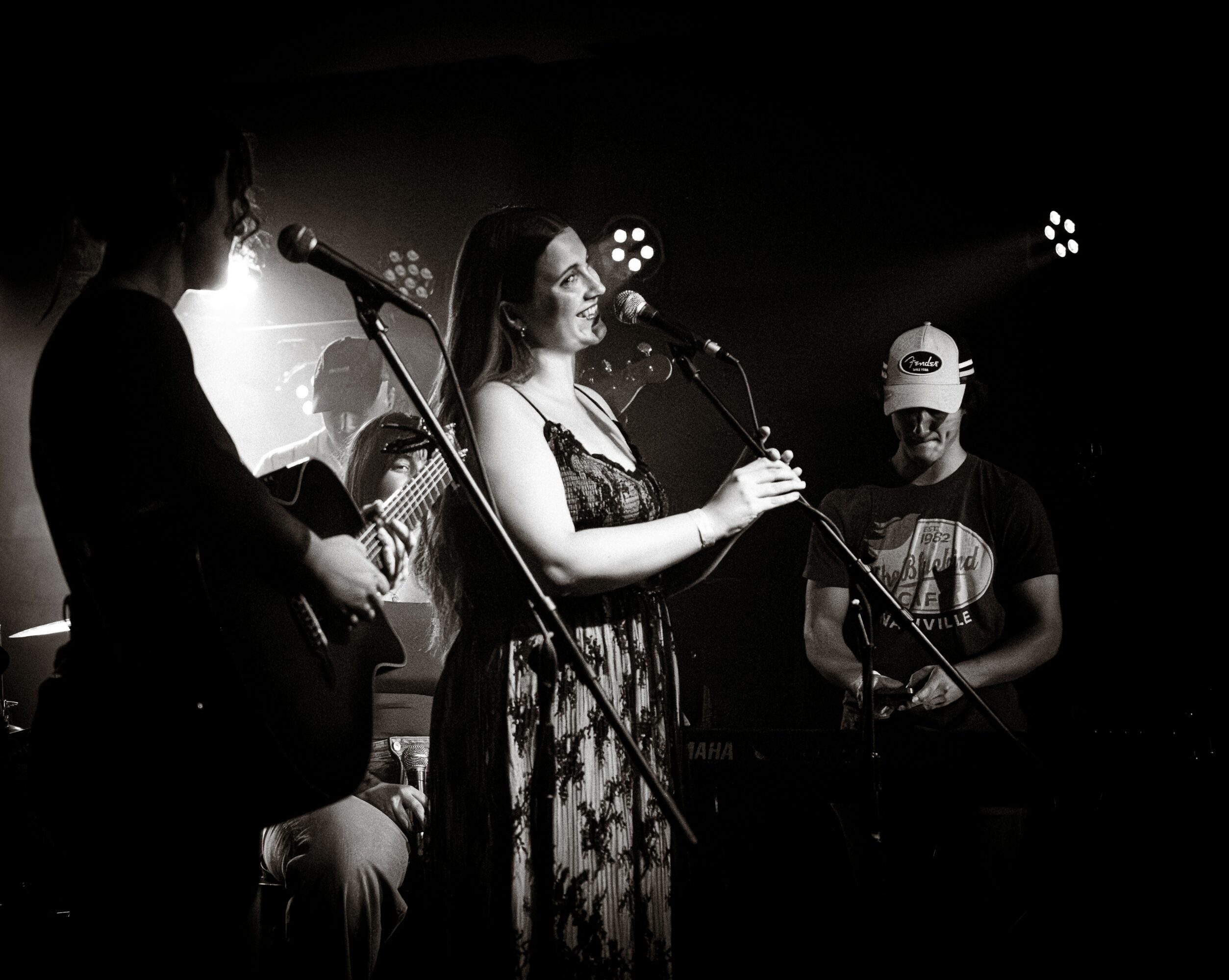 Three musicians perform on stage with microphones and instruments, in black and white, with stage lights in the background.
