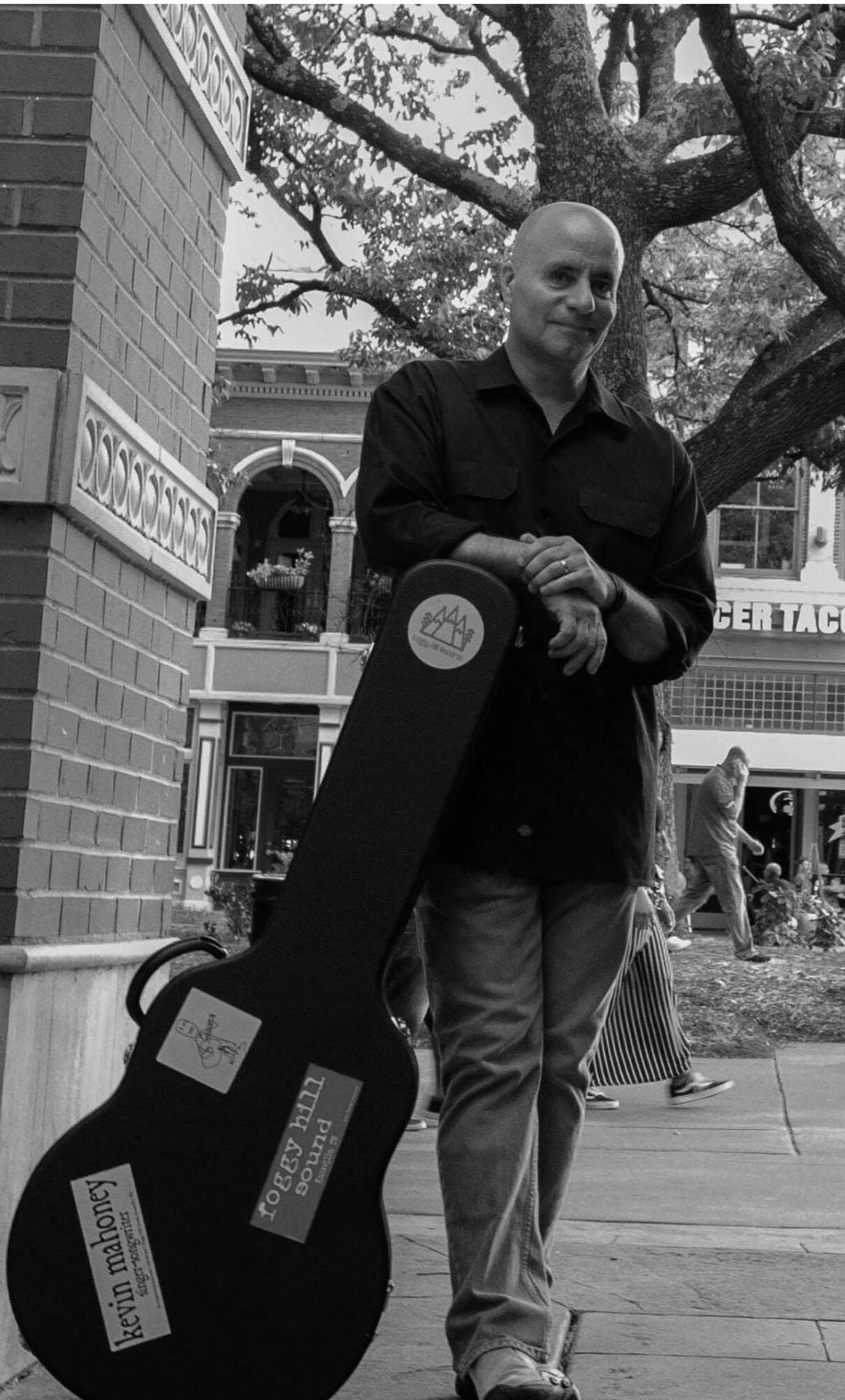 Man standing outdoors holding a guitar case, with a tree and buildings in the background.