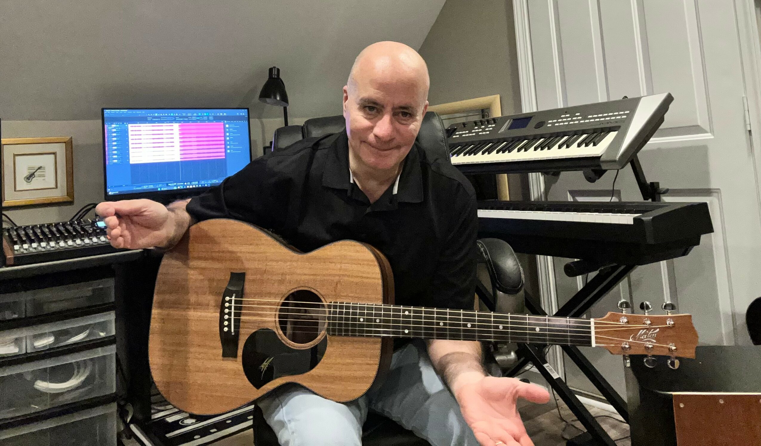 Man holding an acoustic guitar in a music studio with keyboard and computer in background.