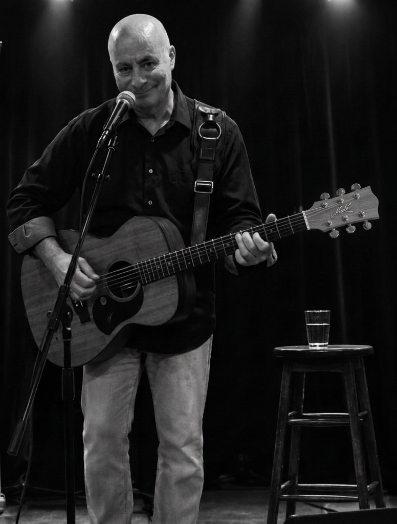 Bald man playing guitar and singing into microphone on stage with a stool and glass of water nearby