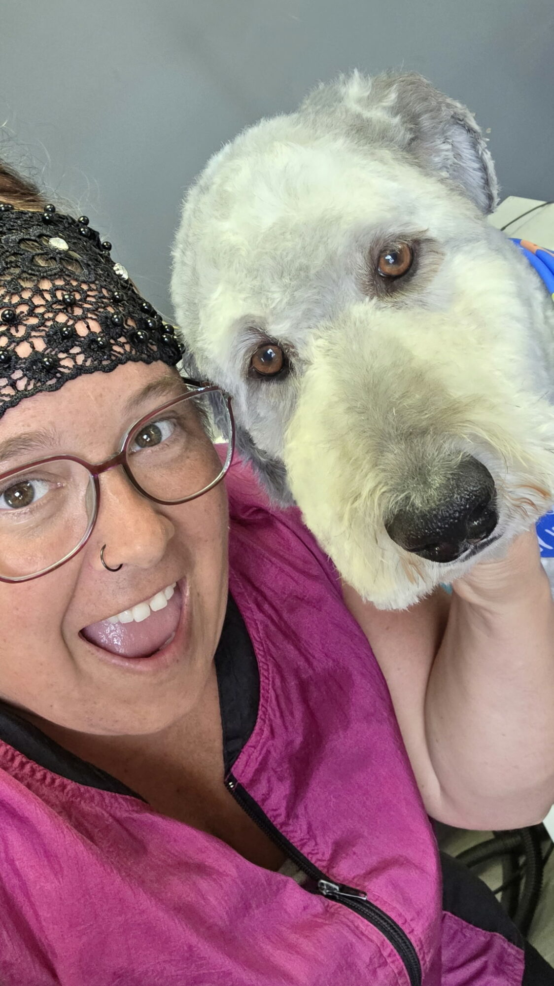 Person with glasses and a nose piercing holding a large white dog close, both looking at the camera.