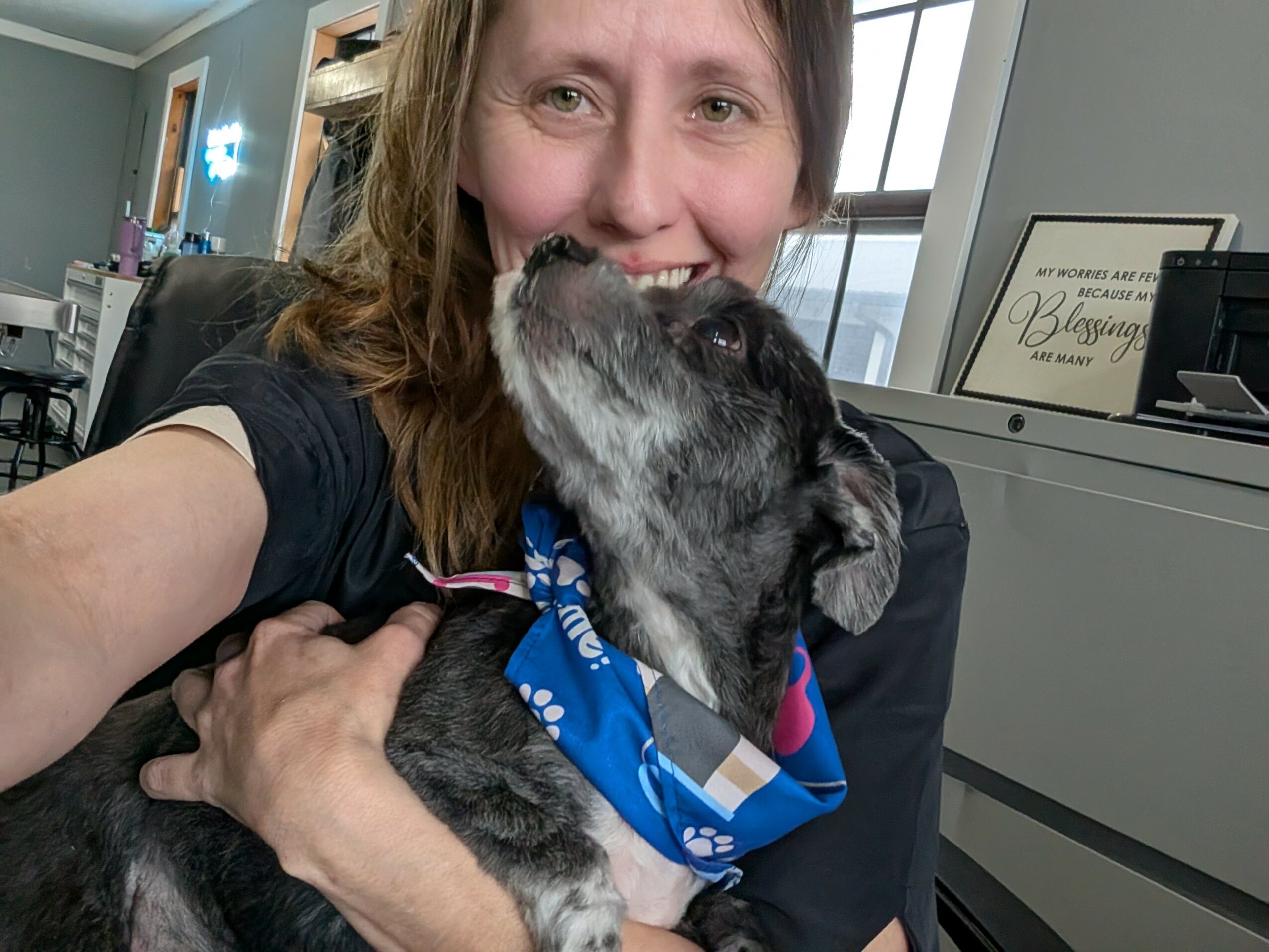 Person smiling with a dog wearing a blue bandana, indoors near a window and desk.
