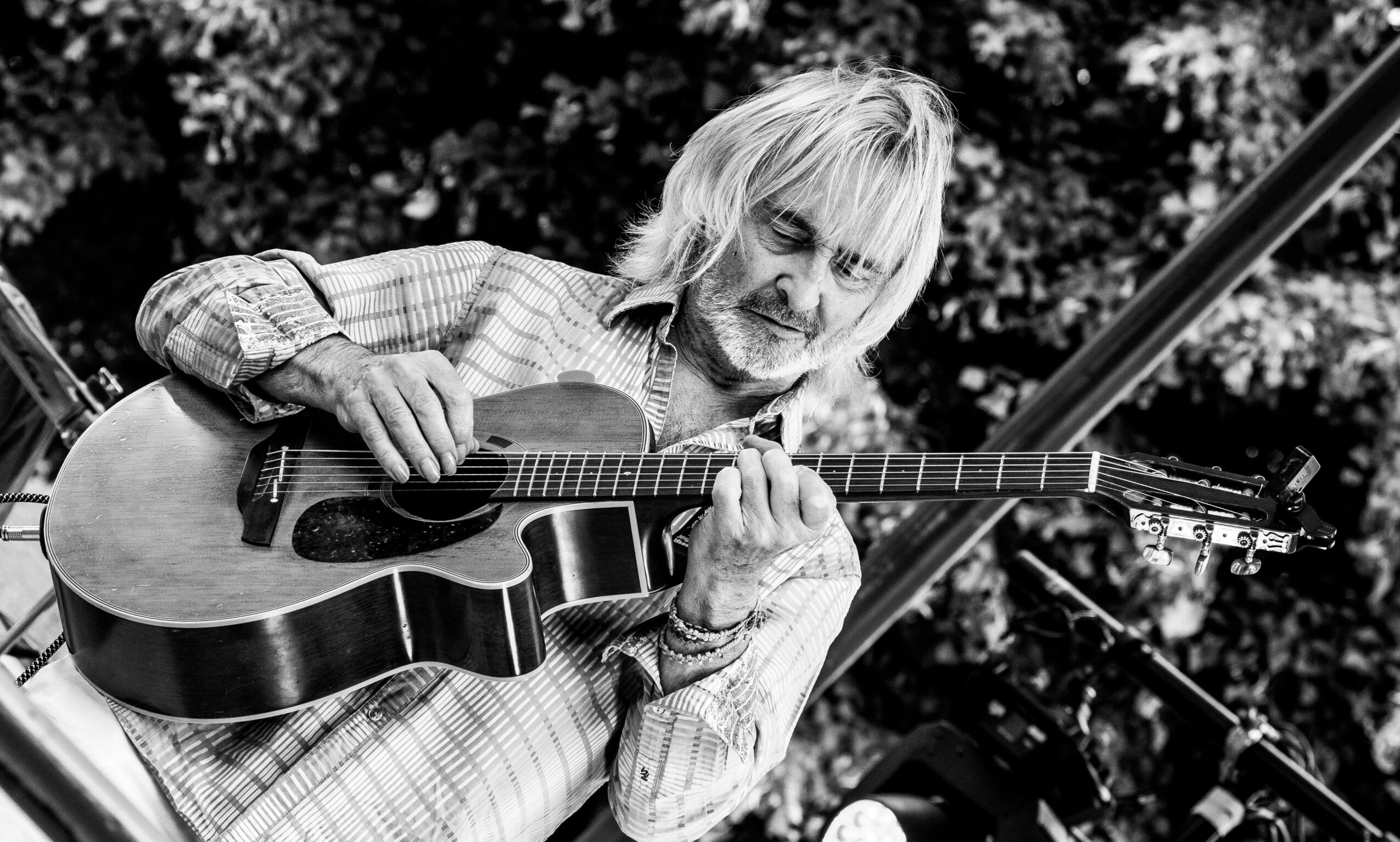 Older man with long hair and beard playing an acoustic guitar outdoors, smiling, with trees in the background.