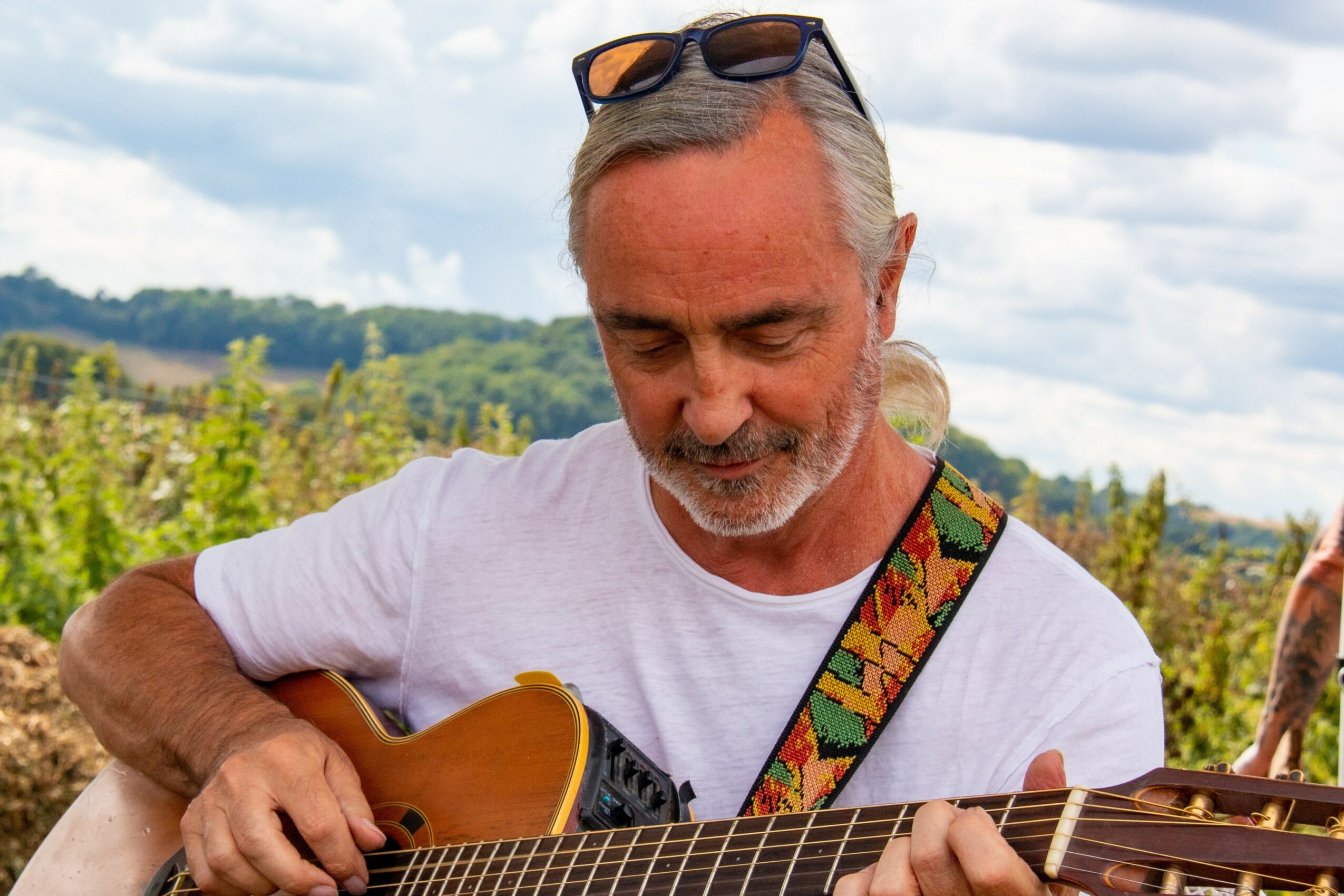 Older man with gray hair and beard playing guitar outdoors, wearing sunglasses on his head and a white shirt.