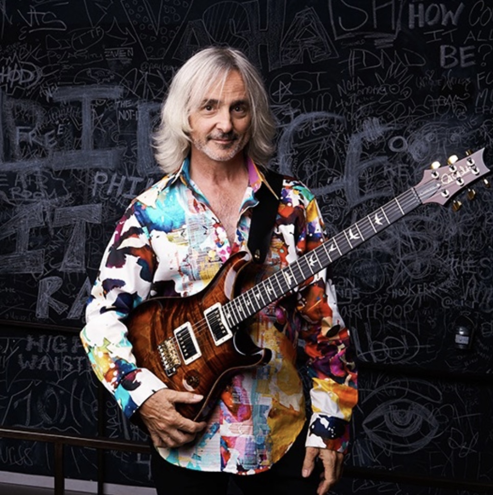 Man with long gray hair holding an electric guitar, wearing a colorful patterned shirt, standing in front of a chalkboard wall.