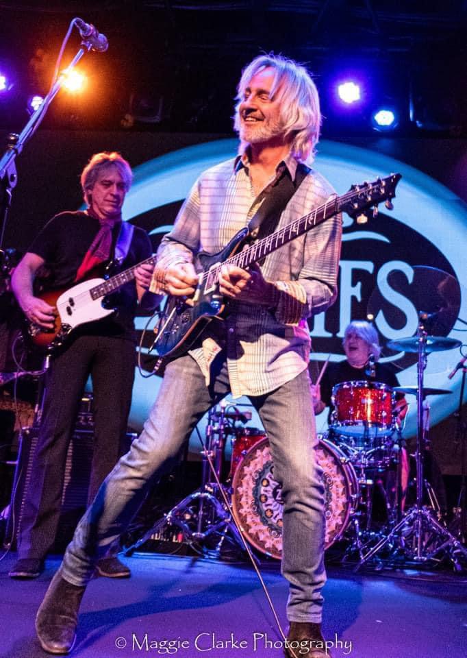 Three musicians perform on stage, with a large circular logo in the background, one playing guitar and singing, others playing instruments.