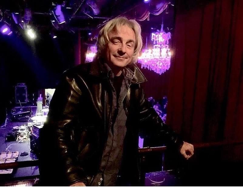 Person with light hair in a leather jacket standing in a stage setting with purple lighting and red curtains.