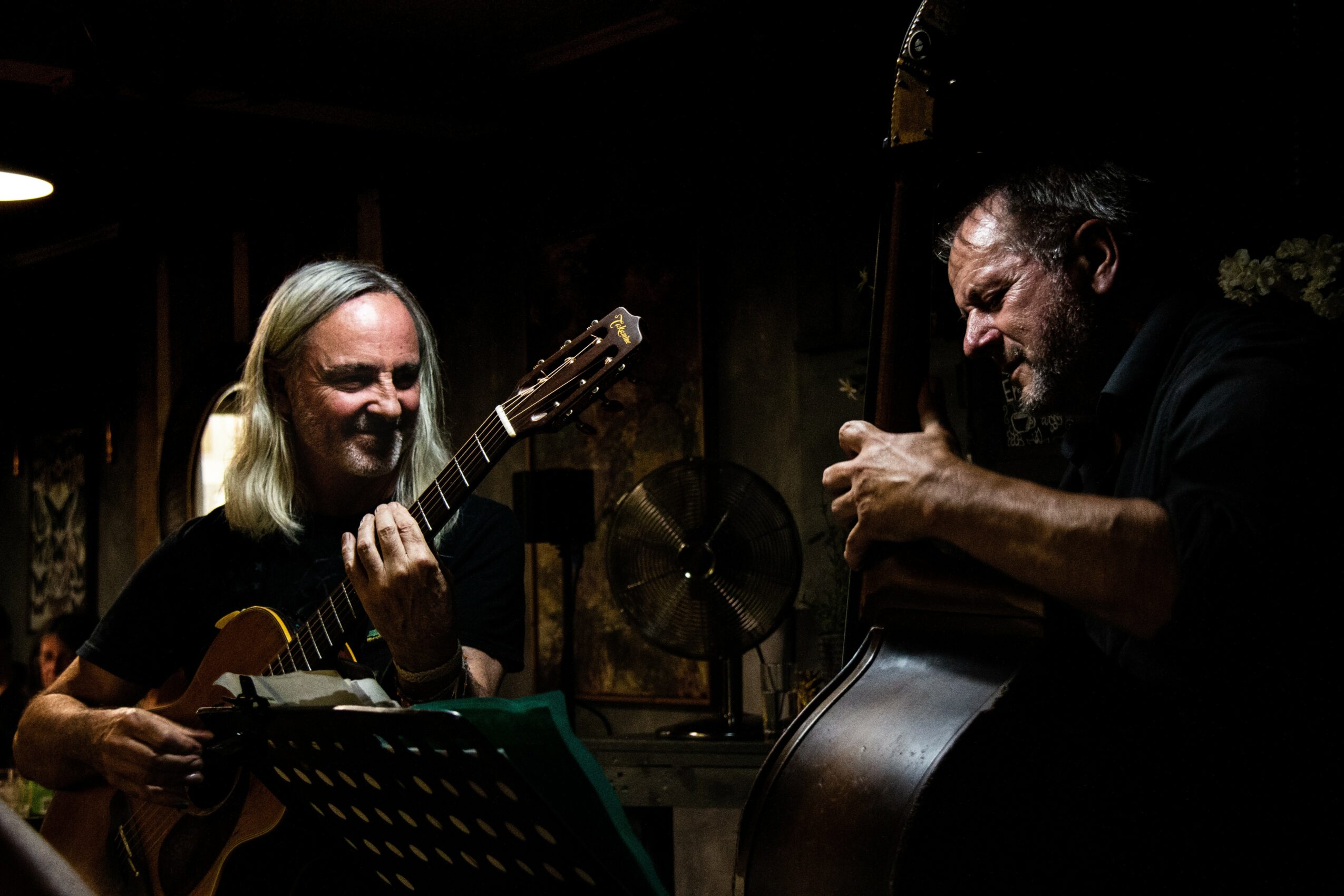 Two people playing guitars in a dimly lit room, smiling and enjoying music together.