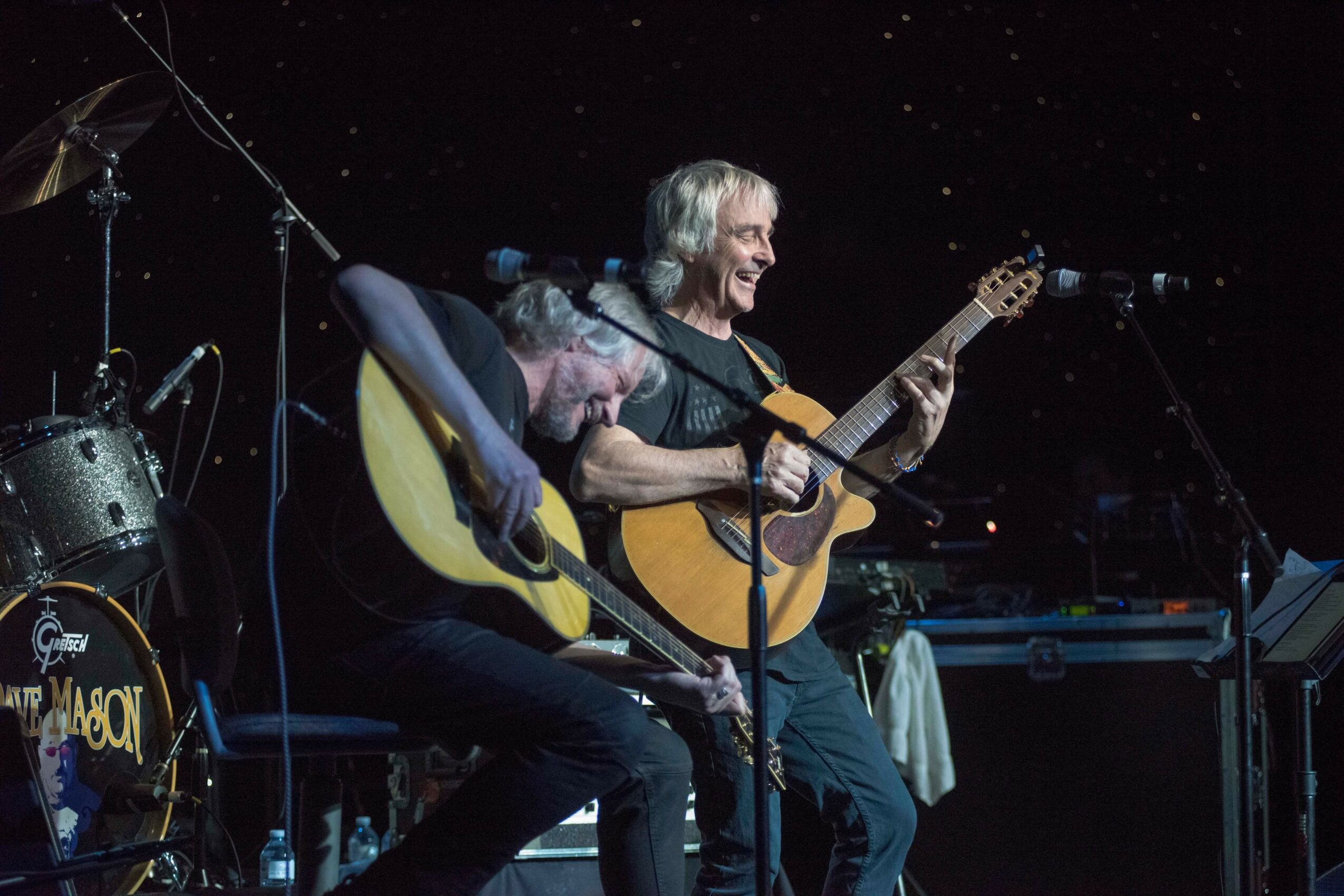 Two musicians playing guitars on stage with microphones, dark background, stage equipment visible.