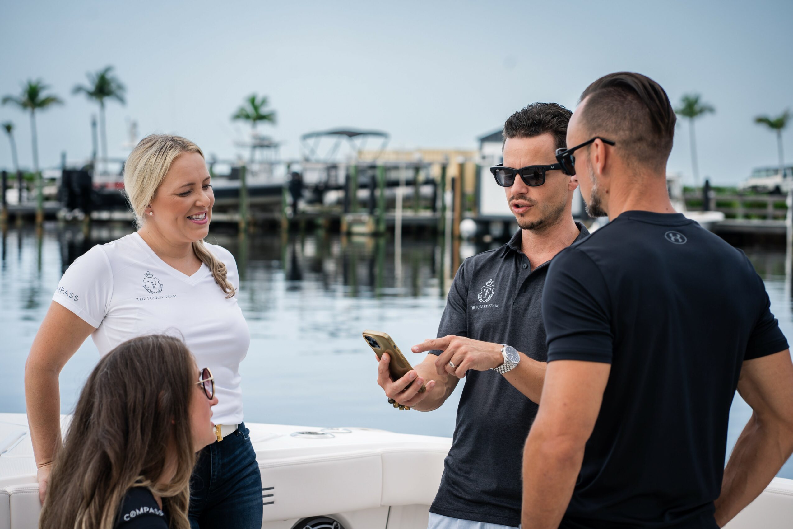 Four people stand near water with boats and palm trees in the background, engaging in conversation outdoors.