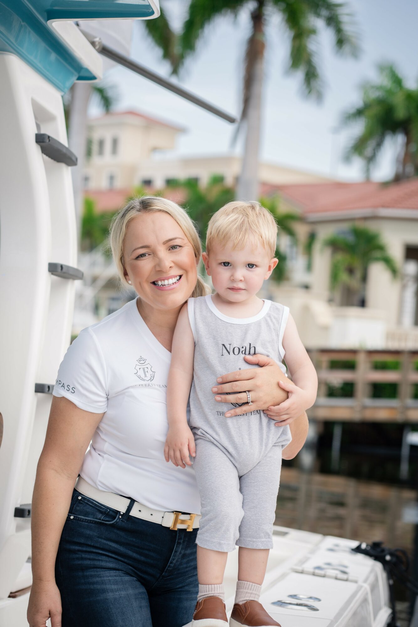 Smiling woman holding a young child outdoors near a dock, with houses and palm trees in the background.