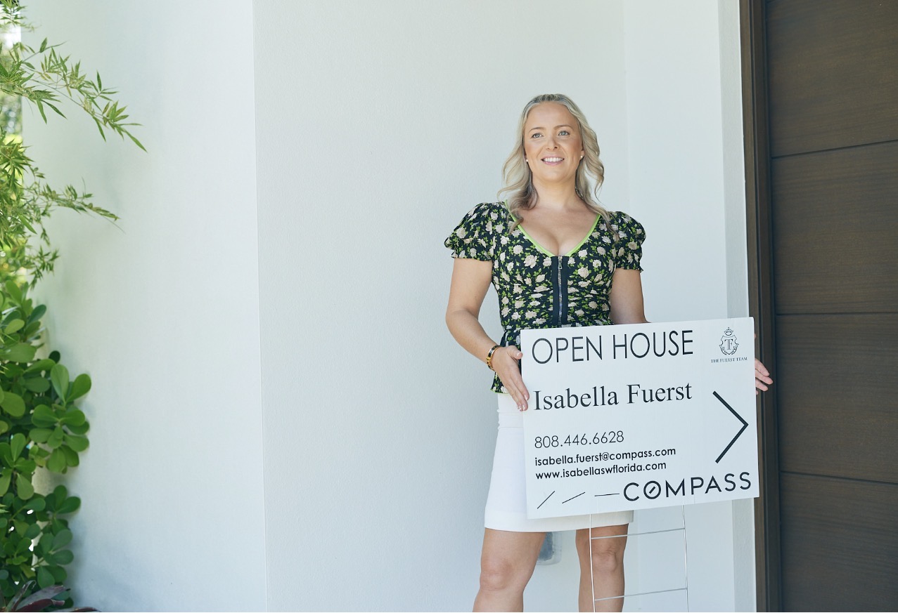 Woman holding an open house sign outside, smiling, with a white wall and dark door in the background.