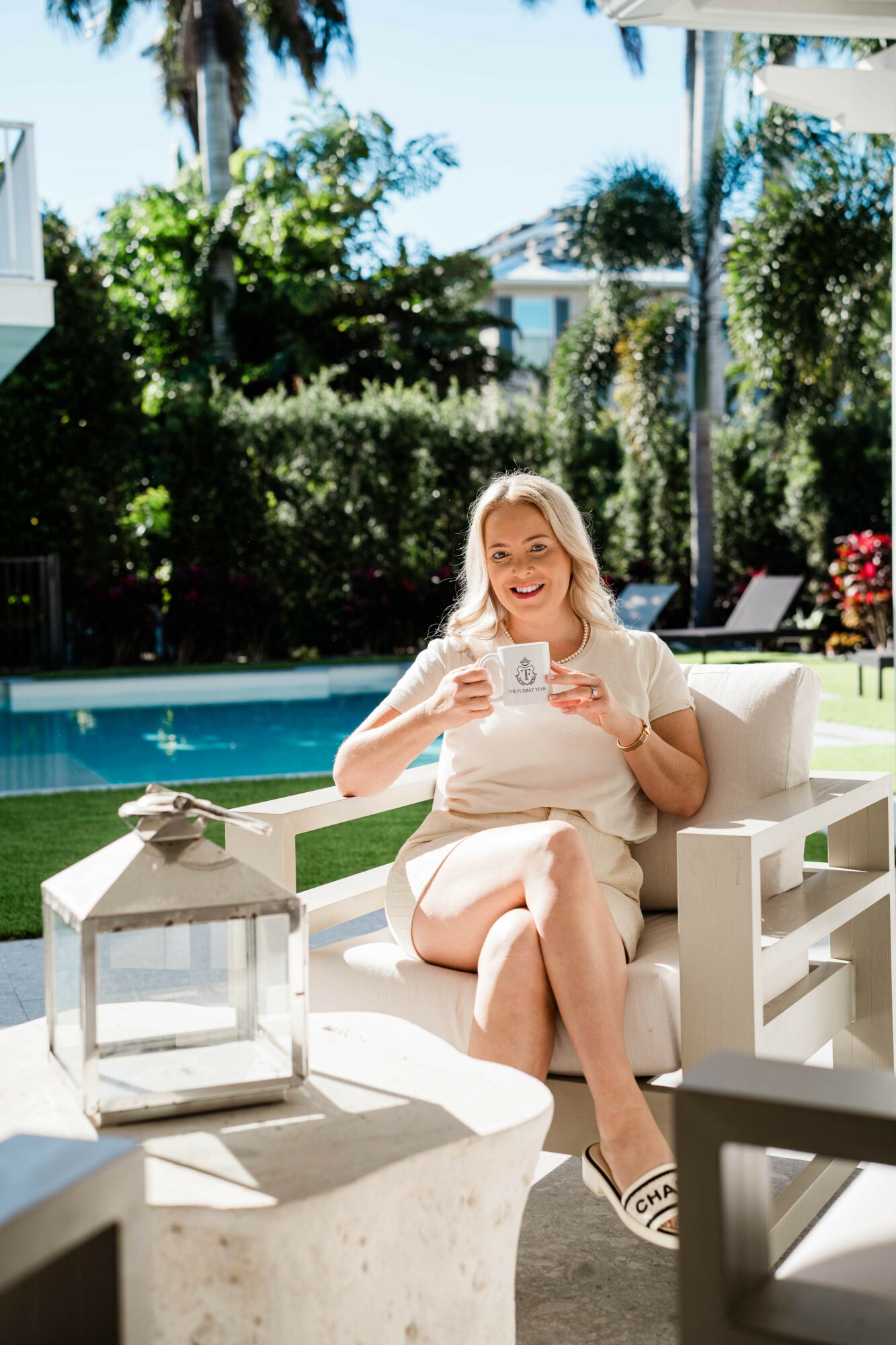 Pregnant woman sitting on a white outdoor chair by a pool, smiling, holding a cup, sunny day with trees and houses in background.