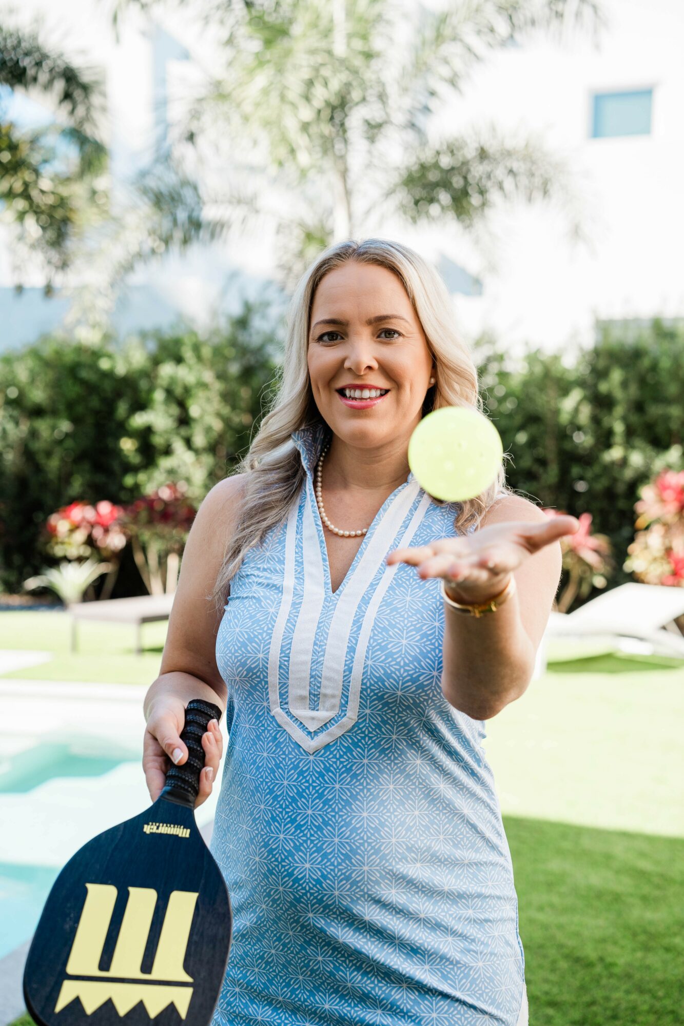 Woman smiling outdoors, holding a tennis ball towards the camera, with a tennis racket in her other hand, in a garden setting.