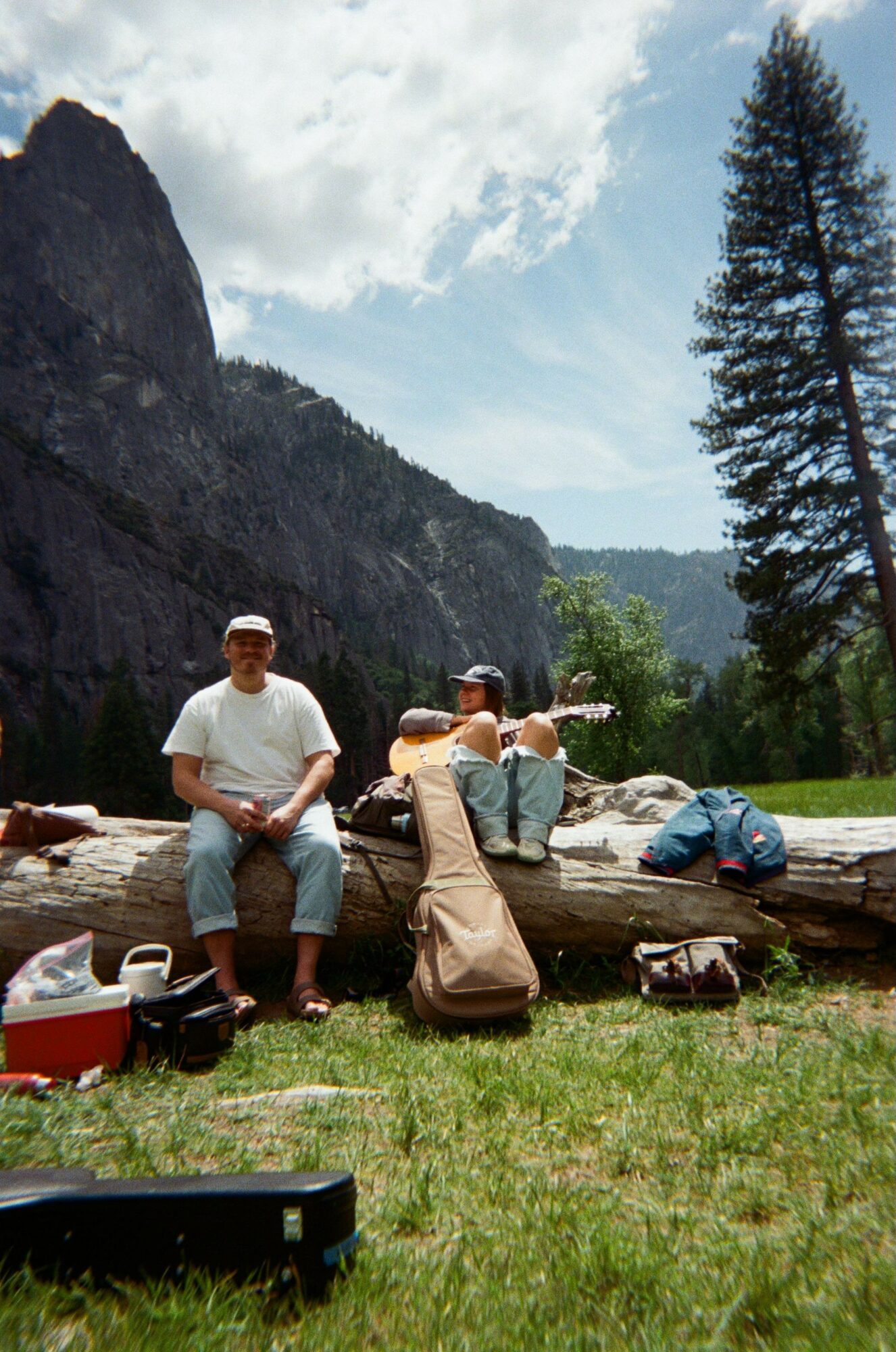 Group of people outdoors near a mountain with trees and cloudy sky, some sitting and some standing, with bags and equipment.