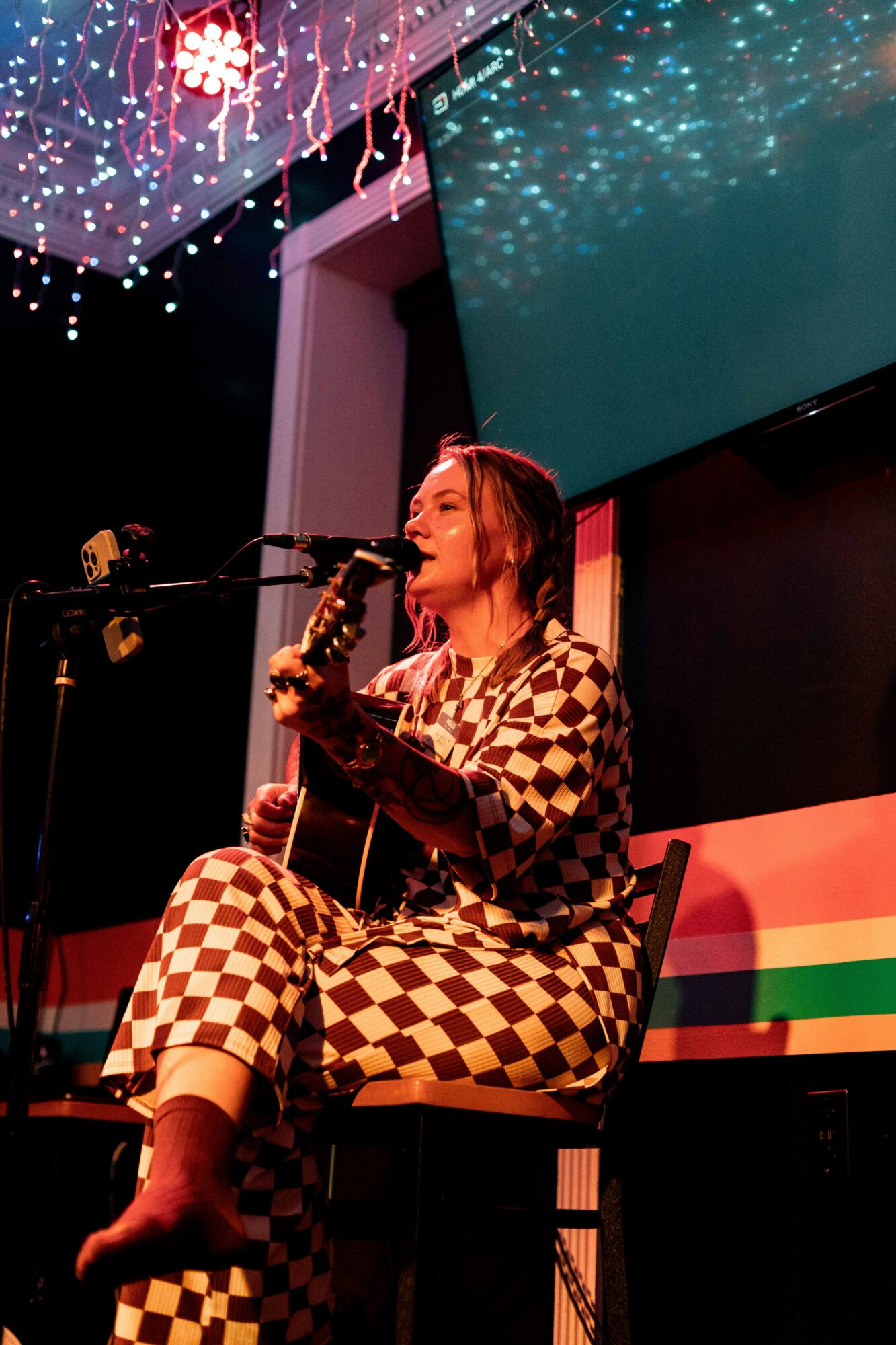 Person sitting on a chair, playing guitar and singing into a microphone, with colorful decorations overhead and a rainbow-colored backdrop.