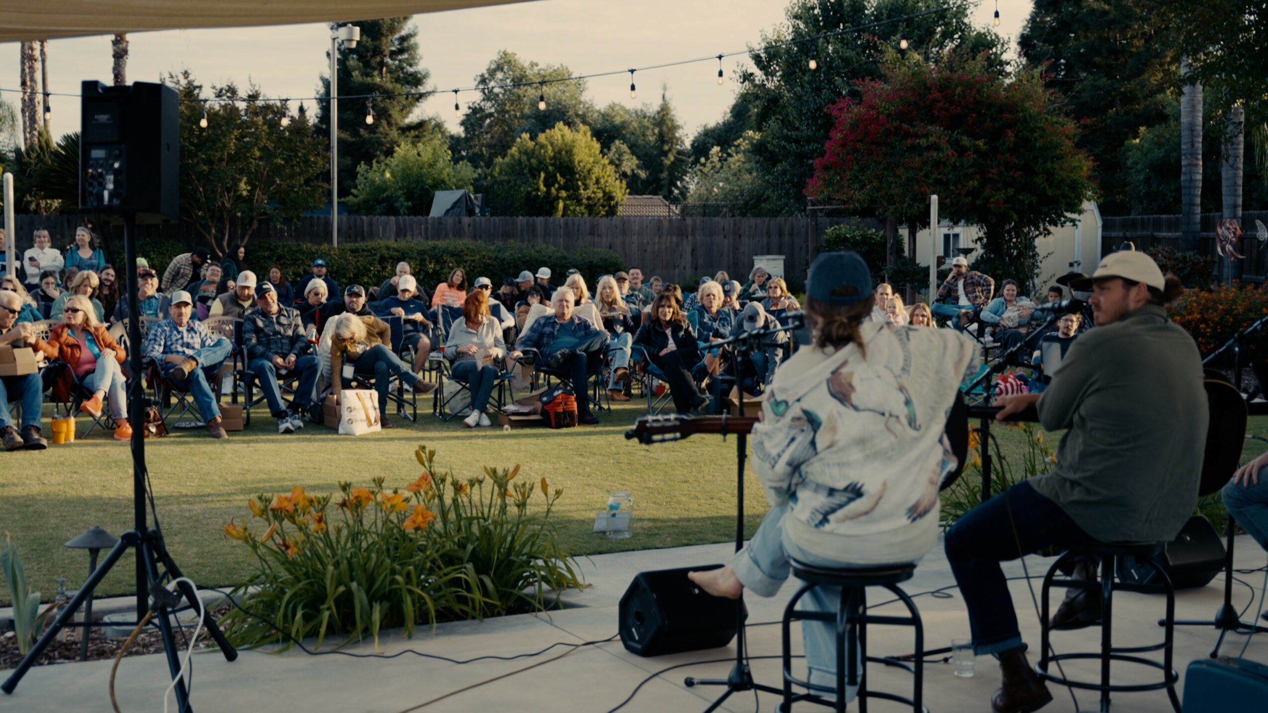 Two people sit on stools on stage, facing an outdoor audience seated on chairs, with trees and a fence in background.