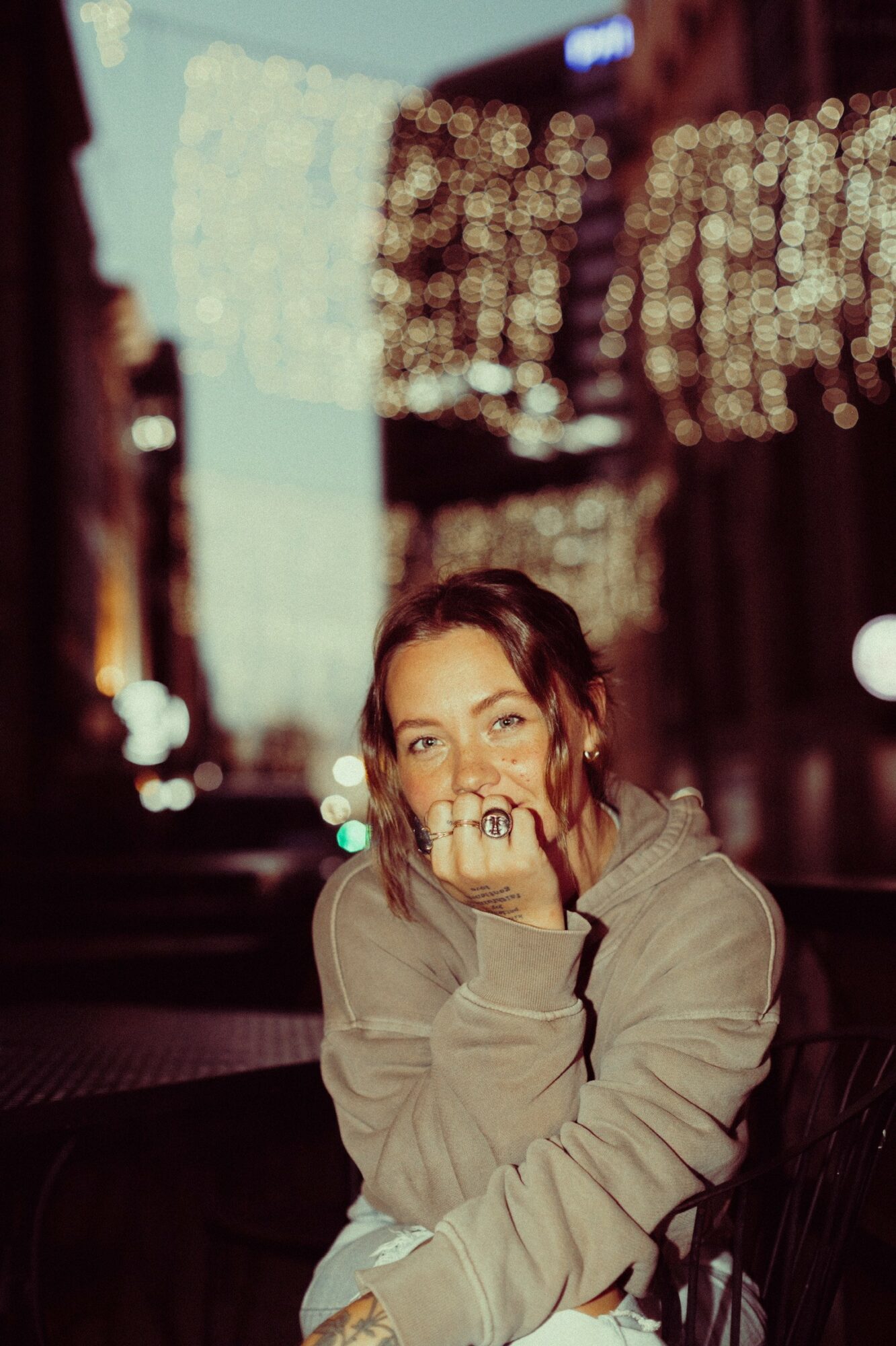Young woman with dark hair and a beige hoodie, sitting outdoors at night with city lights in the background.