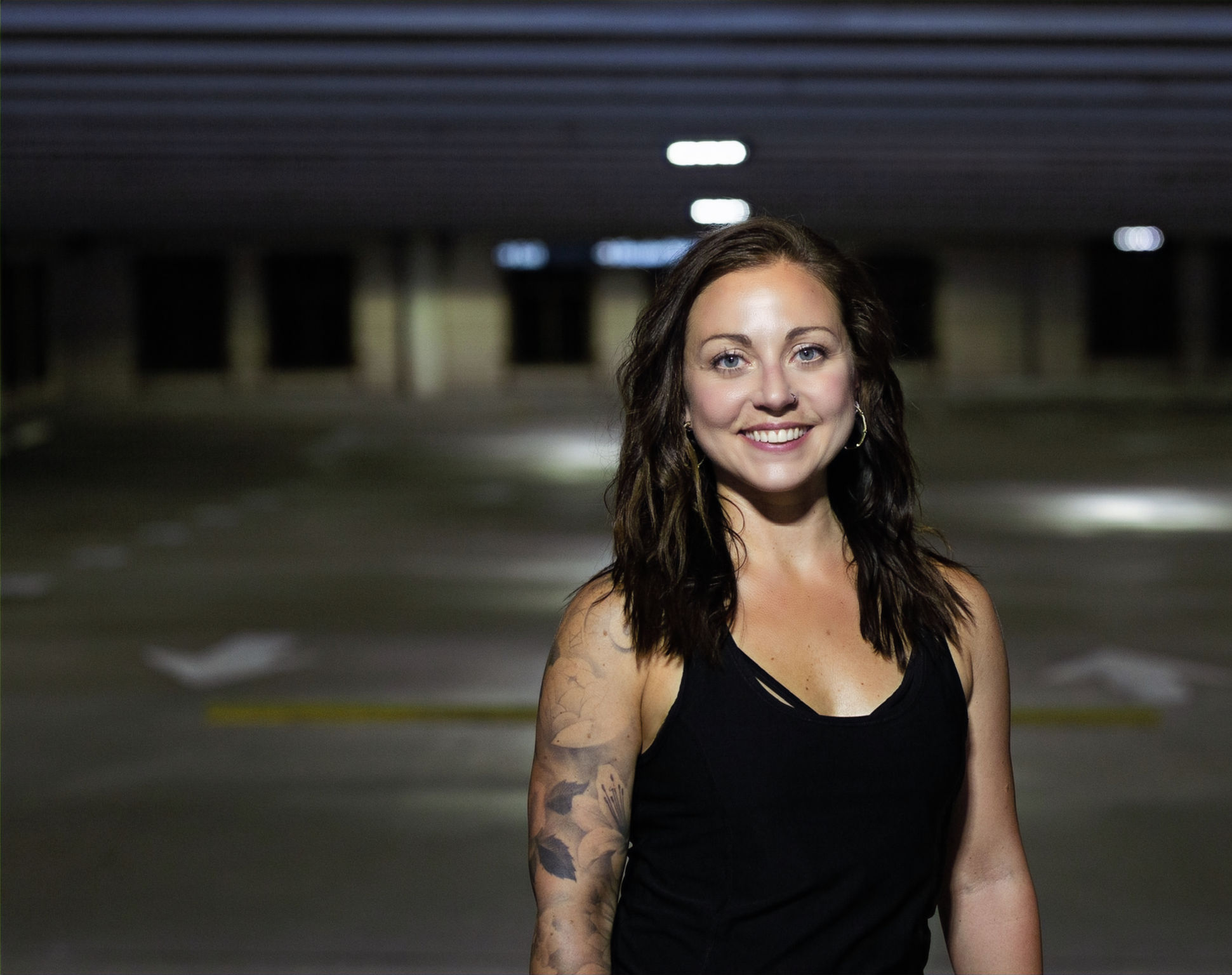 Young woman with dark hair and tattoos smiling in an empty parking garage, wearing a black top.