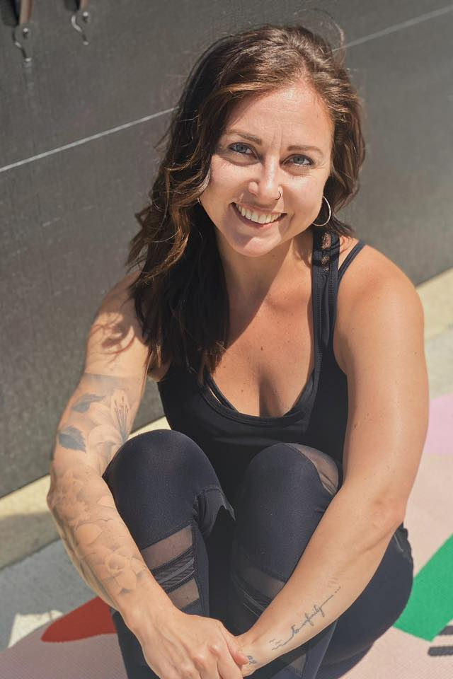 Smiling woman with long dark hair, wearing a black tank top, sitting outdoors with a dark wall background.