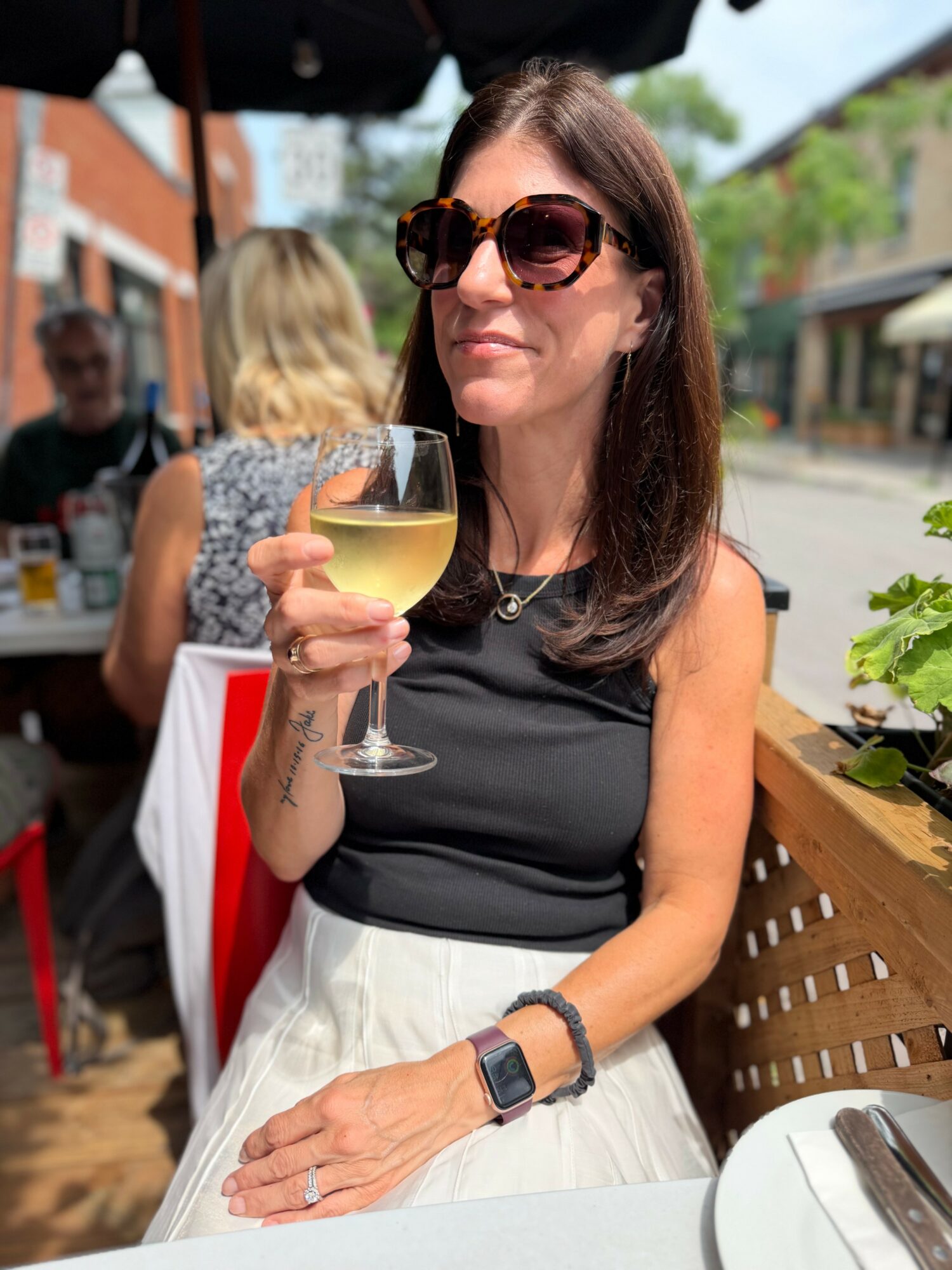 Woman with sunglasses holding a glass of white wine outdoors, wearing a black top and white skirt, smiling.