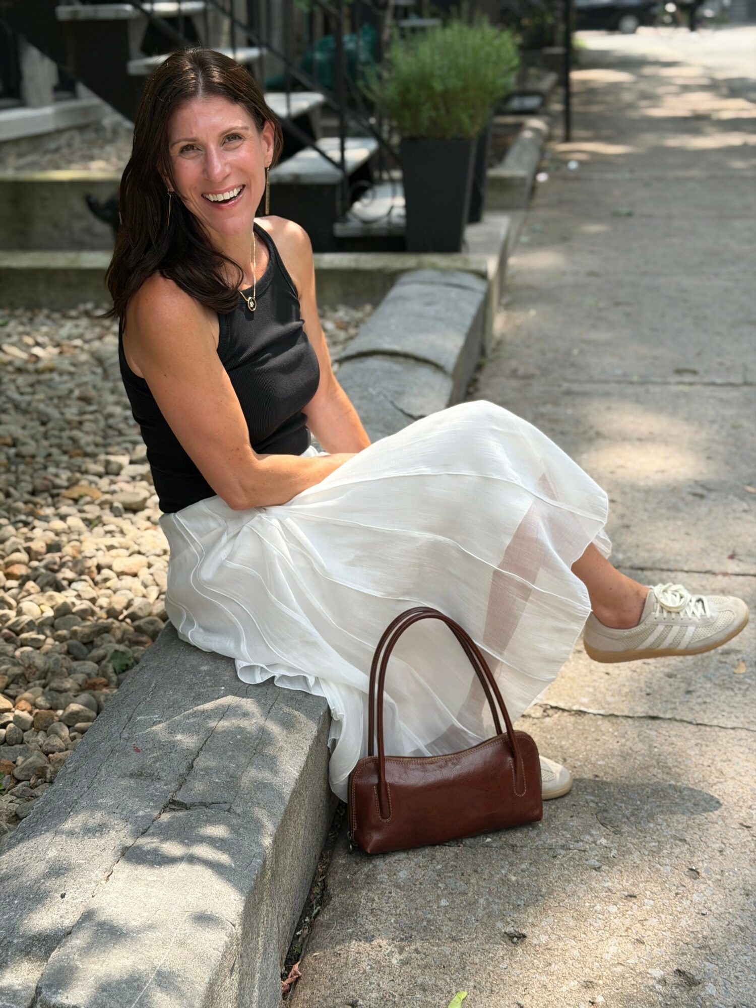 Woman sitting on a curb, smiling, wearing a black top and white skirt, with a brown handbag nearby.