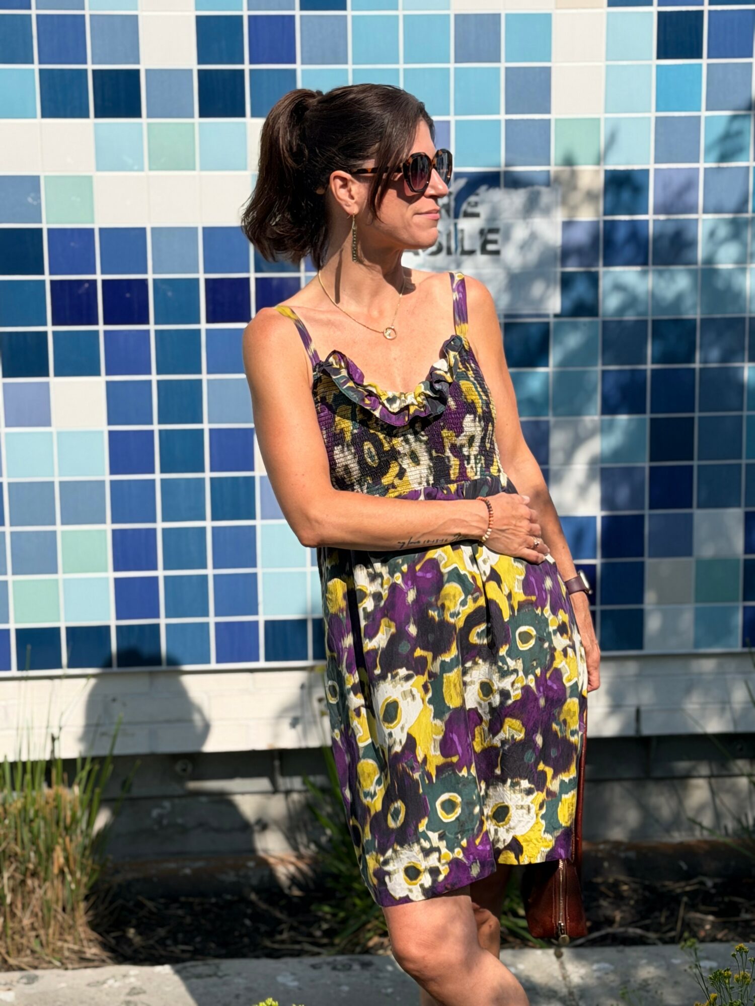 Woman in sunglasses and a colorful floral dress stands outdoors in front of a blue tiled wall.