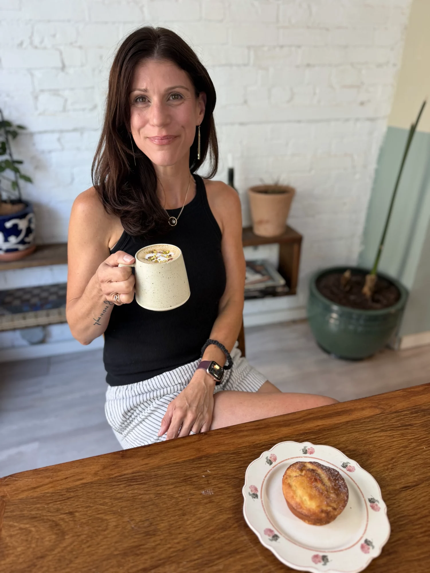 Woman sitting at a table holding a mug, with a plate of pastry in front, in a cozy room with plants and a white brick wall.