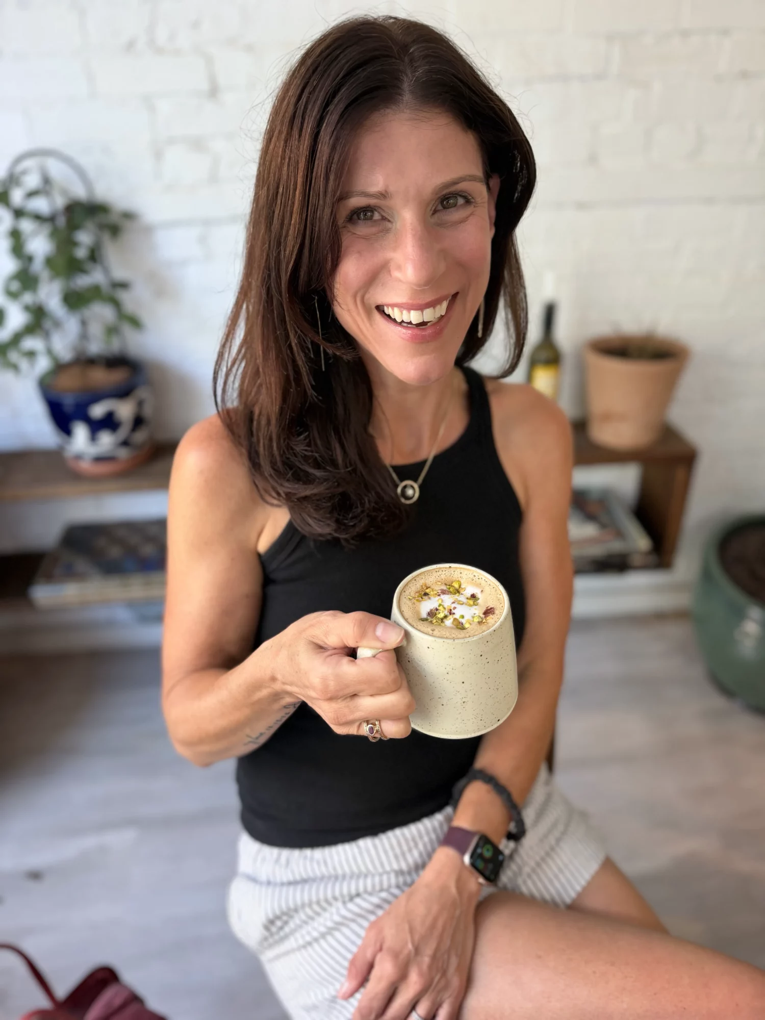 Woman smiling, holding a mug, sitting indoors with a white brick wall and plants in the background.