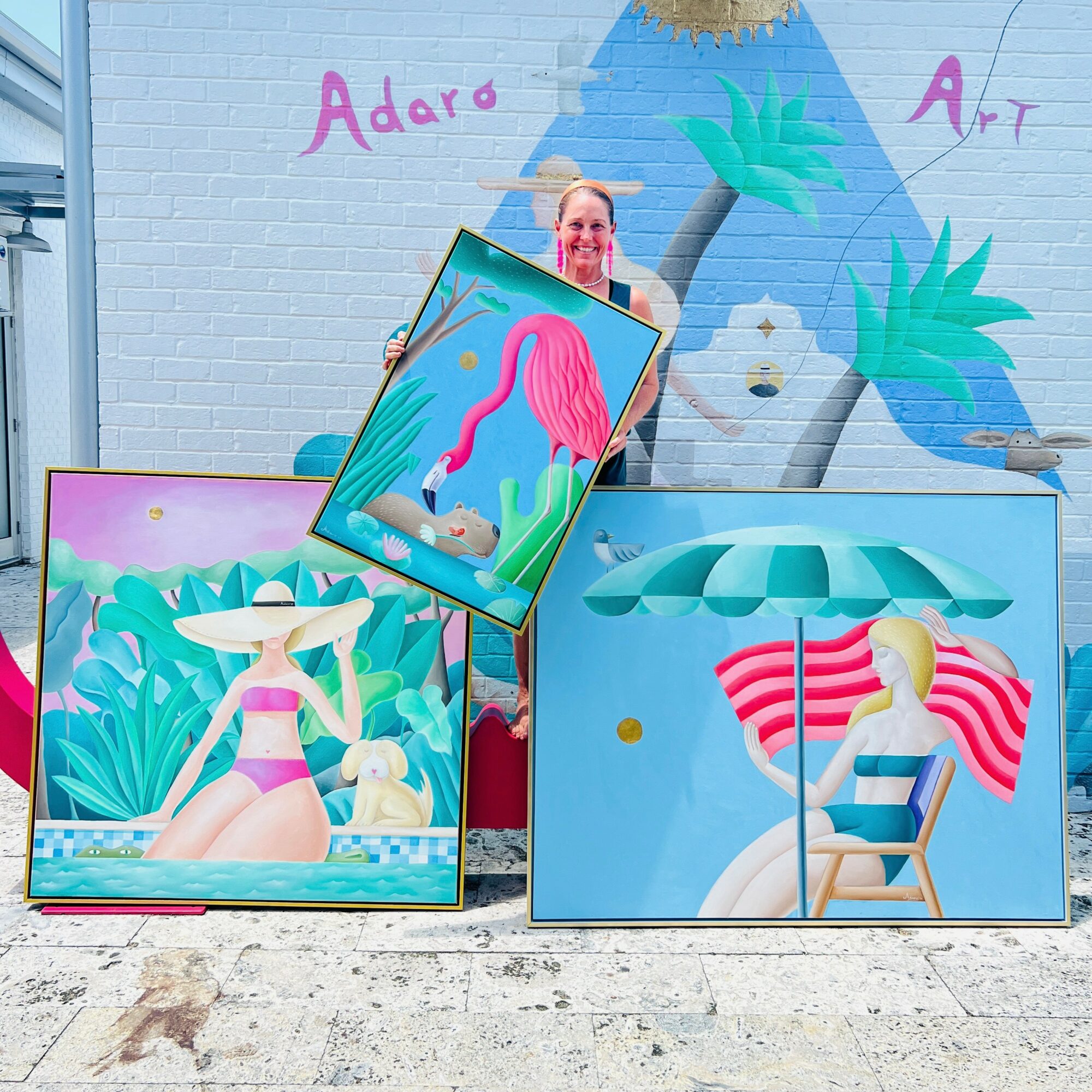 Person holding a framed artwork of a pink flamingo with a blue background, standing outdoors against a colorful mural wall.