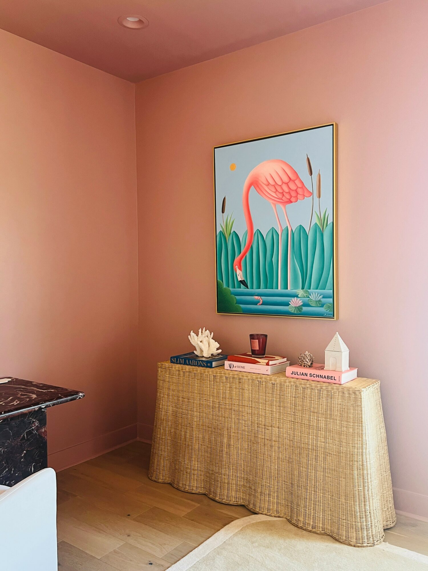 Pink flamingo painting hangs above a table with decorative items, against a pink wall in a room.