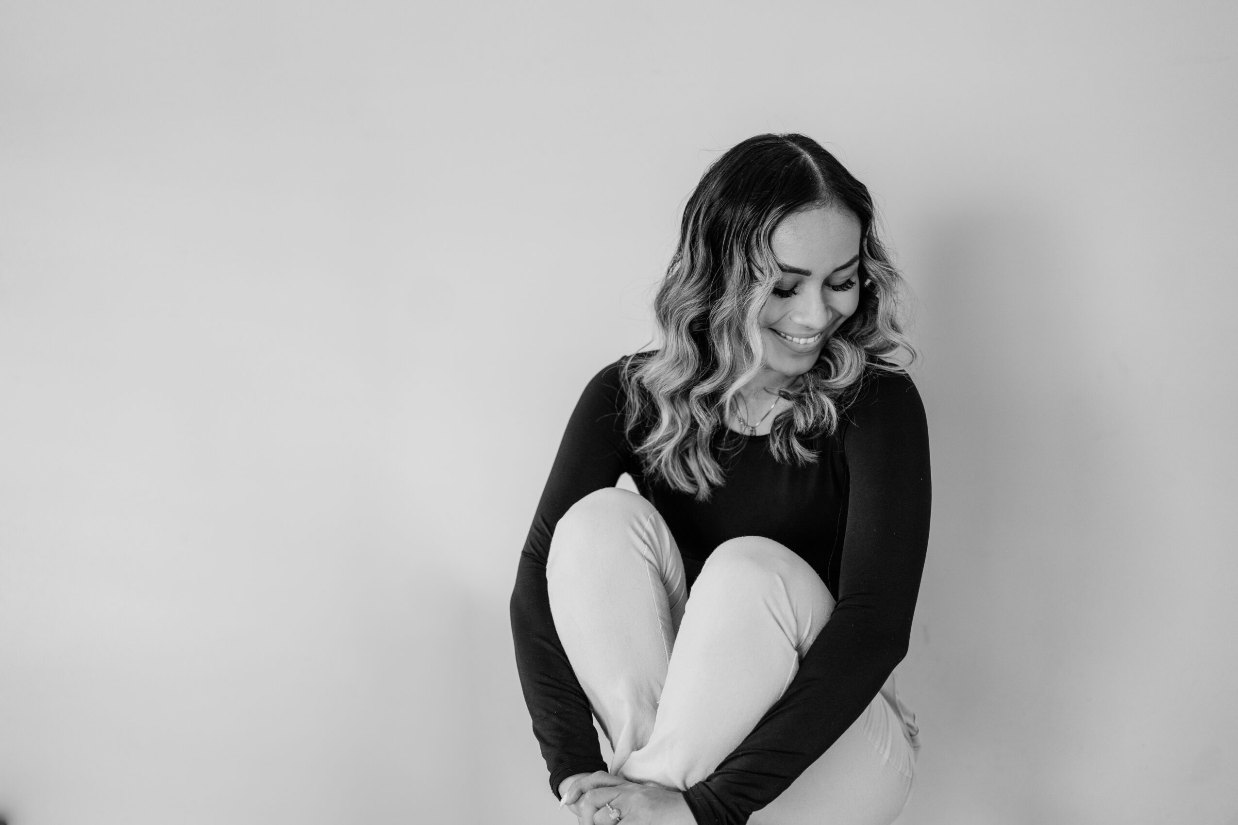 A woman with wavy hair smiling, sitting with her knees up, wearing a black top and light-colored pants, against a plain background.