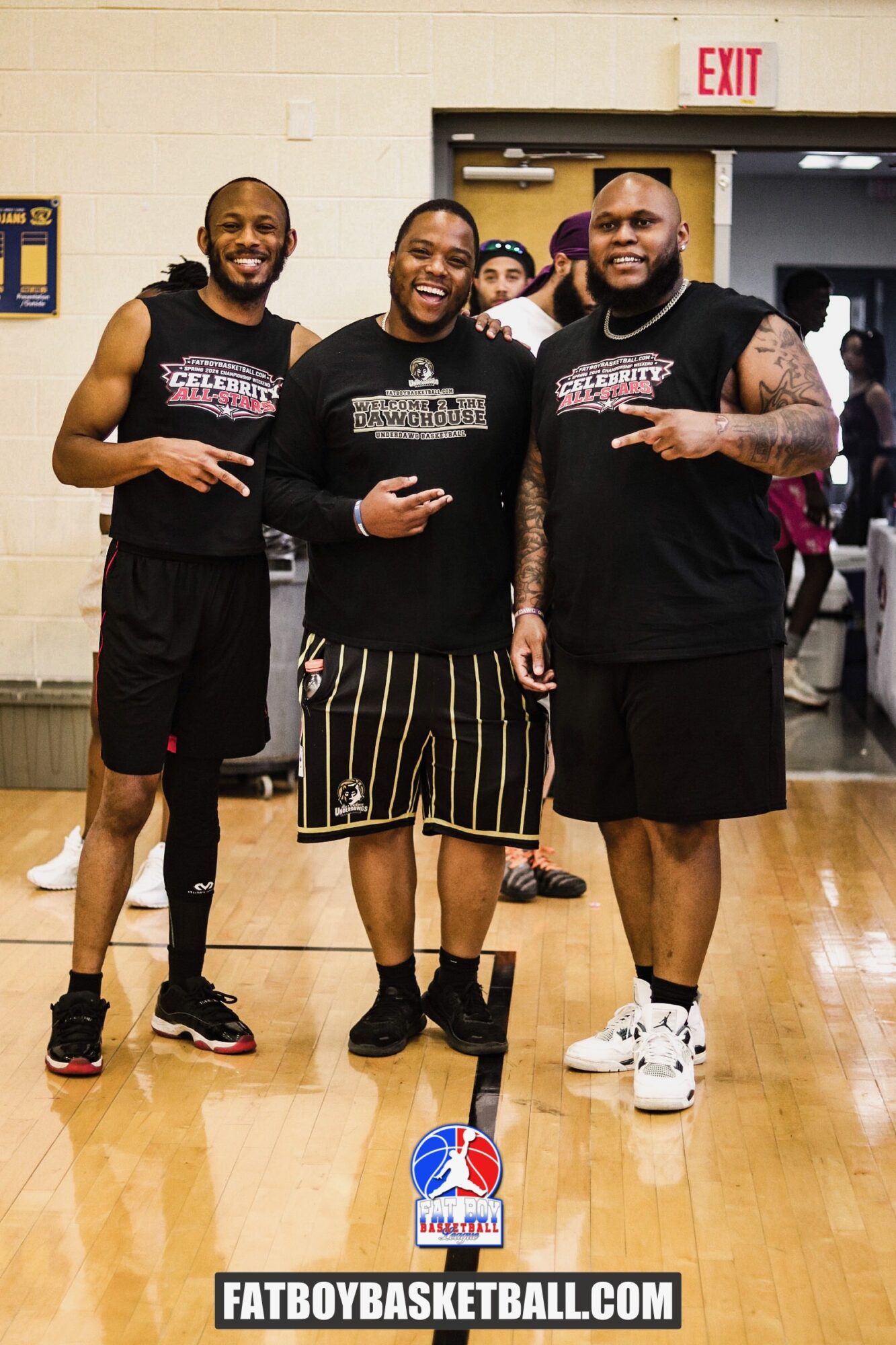 Three basketball players standing indoors, smiling, with a white wall and exit sign behind them.