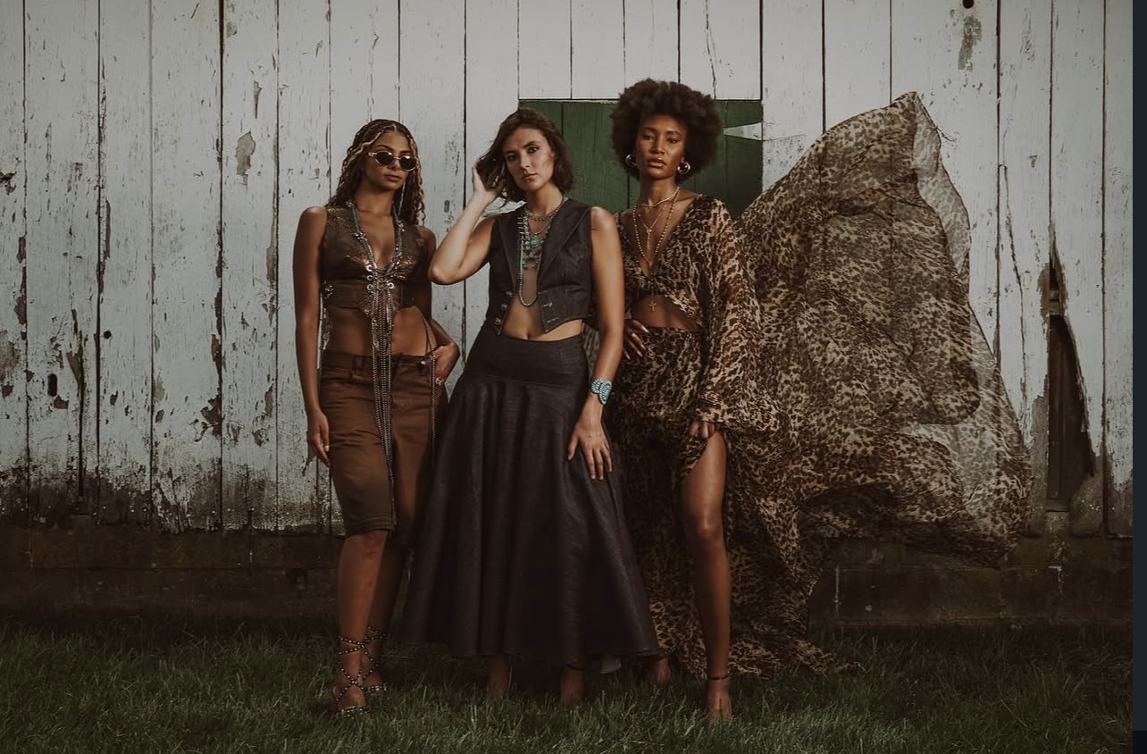 Four women stand outdoors in front of a weathered wooden wall and large rock, dressed in stylish clothing.