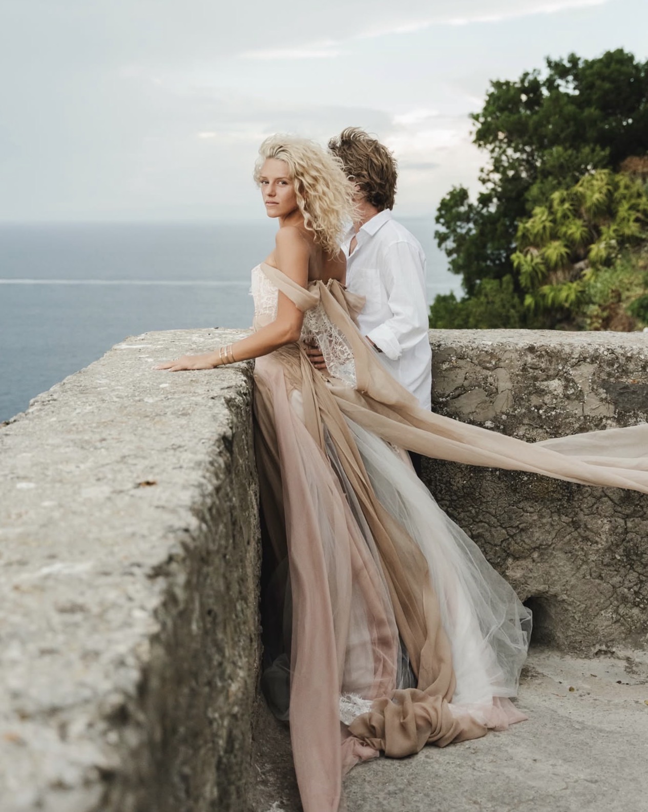 Two people in elegant clothing stand on a stone wall overlooking the sea, with trees in the background.