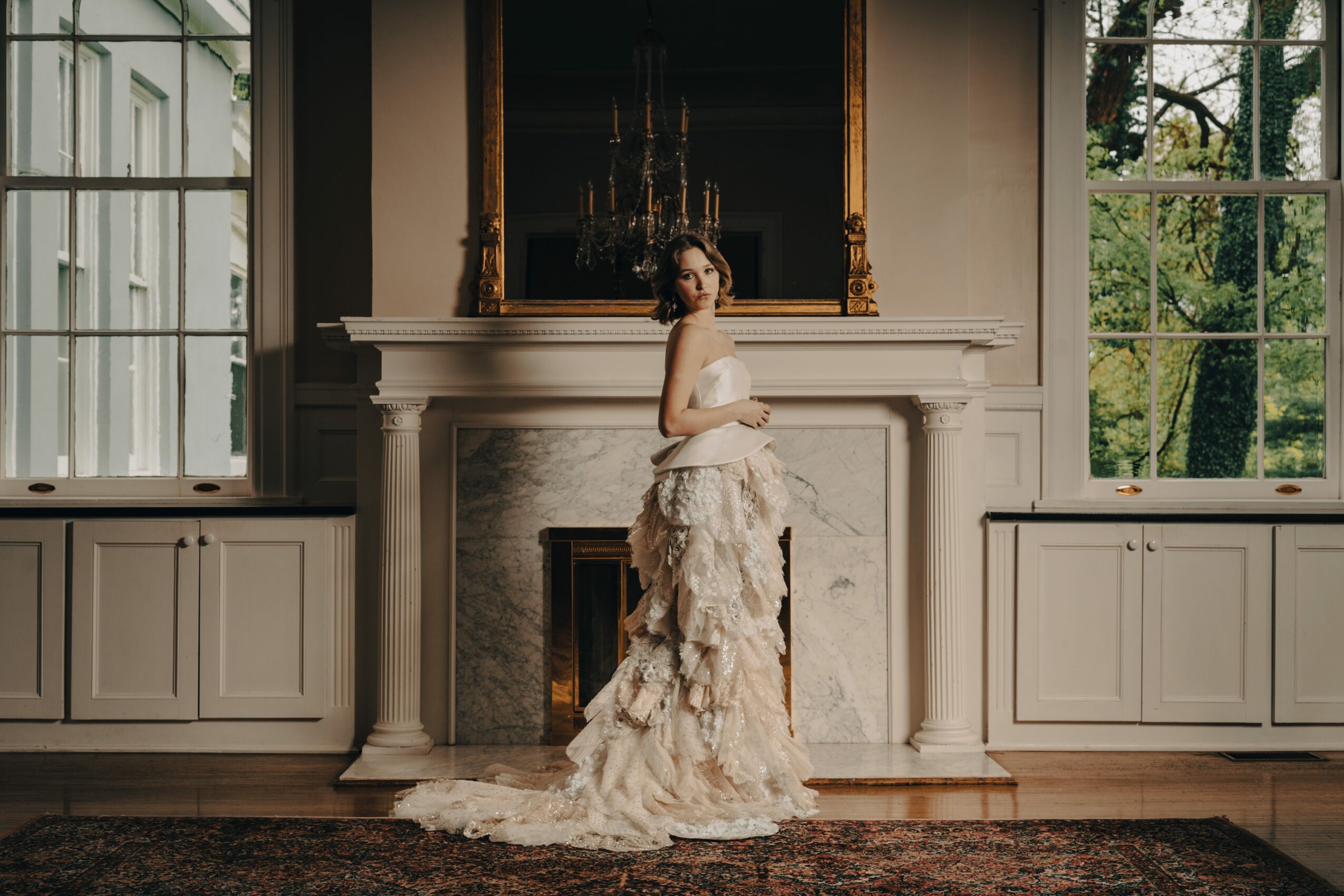 Woman in a wedding dress standing in front of a fireplace in a room with large windows and a mirror above the mantelpiece.