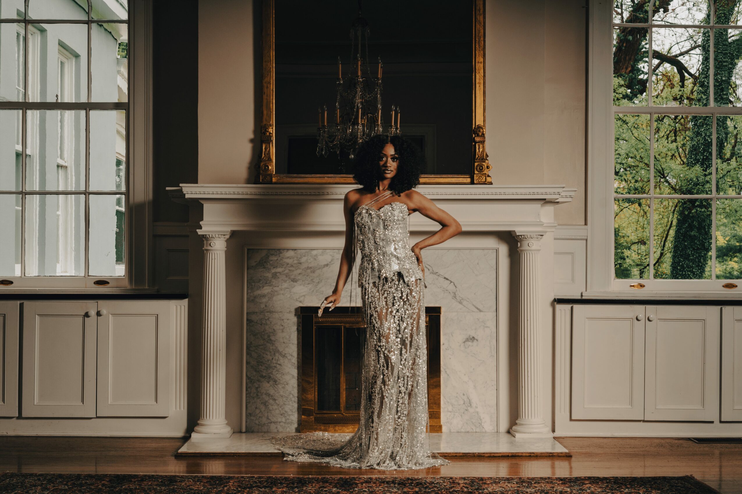 Woman in a long dress standing in front of a fireplace in a room with large windows and a mirror above the fireplace.