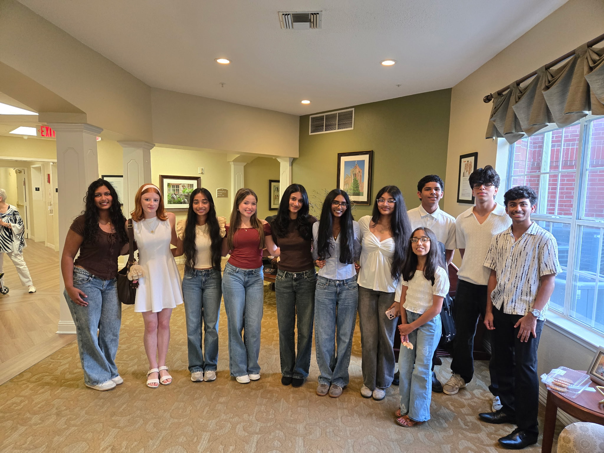 Group of ten people standing indoors, smiling, with windows and framed pictures in the background.