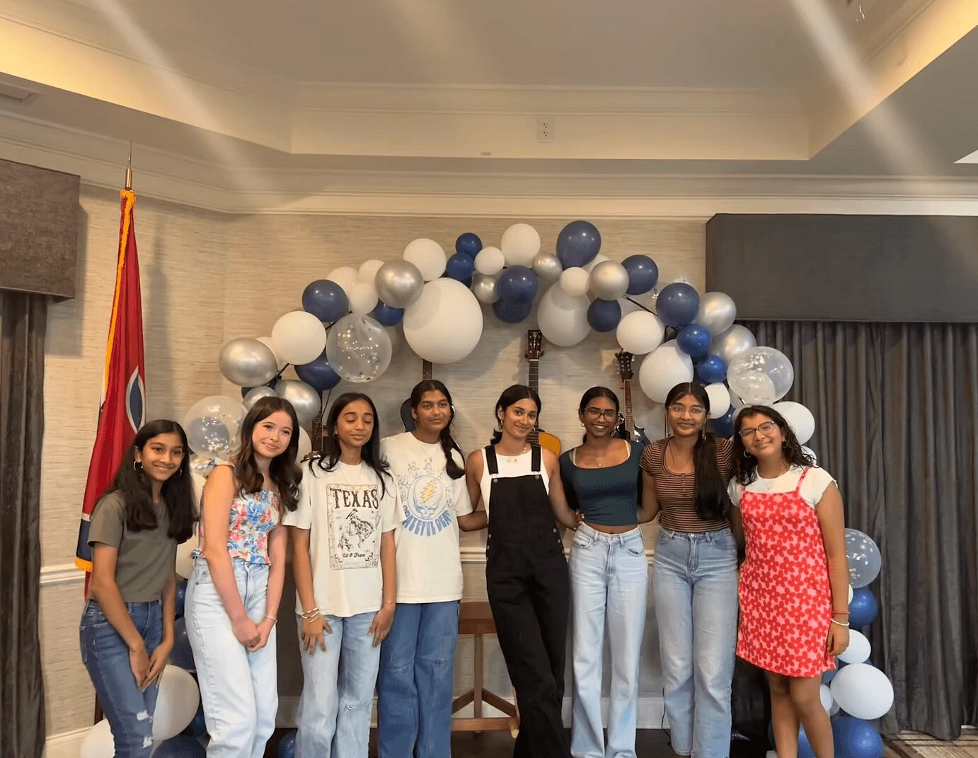 Group of eight women standing in front of balloon arch and flag, smiling, indoors.