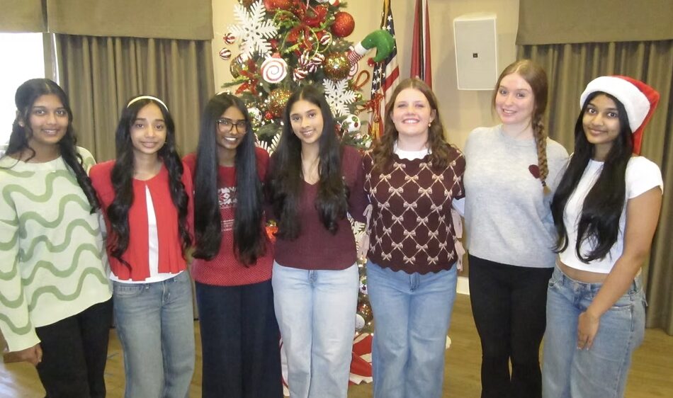 Group of seven young women standing in front of a decorated Christmas tree, smiling, some wearing holiday accessories.